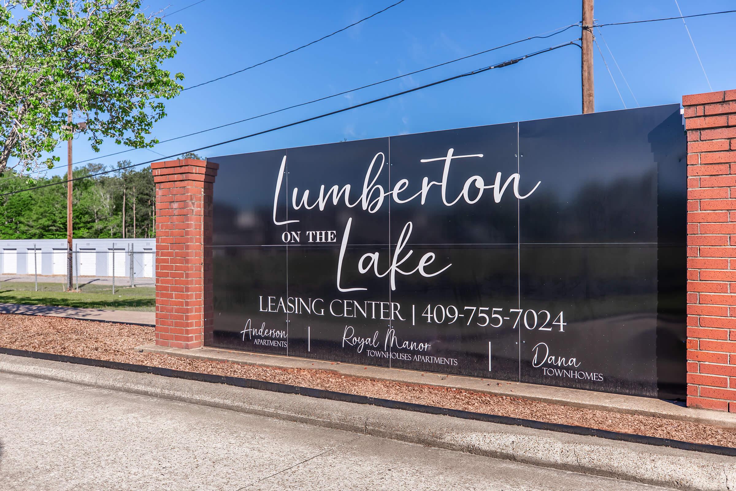 Sign for "Lumberton on the Lake" Leasing Center, featuring contact number 409-755-7024. The sign has a dark background with white script text and is flanked by brick columns. Surrounding greenery is visible, along with a clear blue sky above.