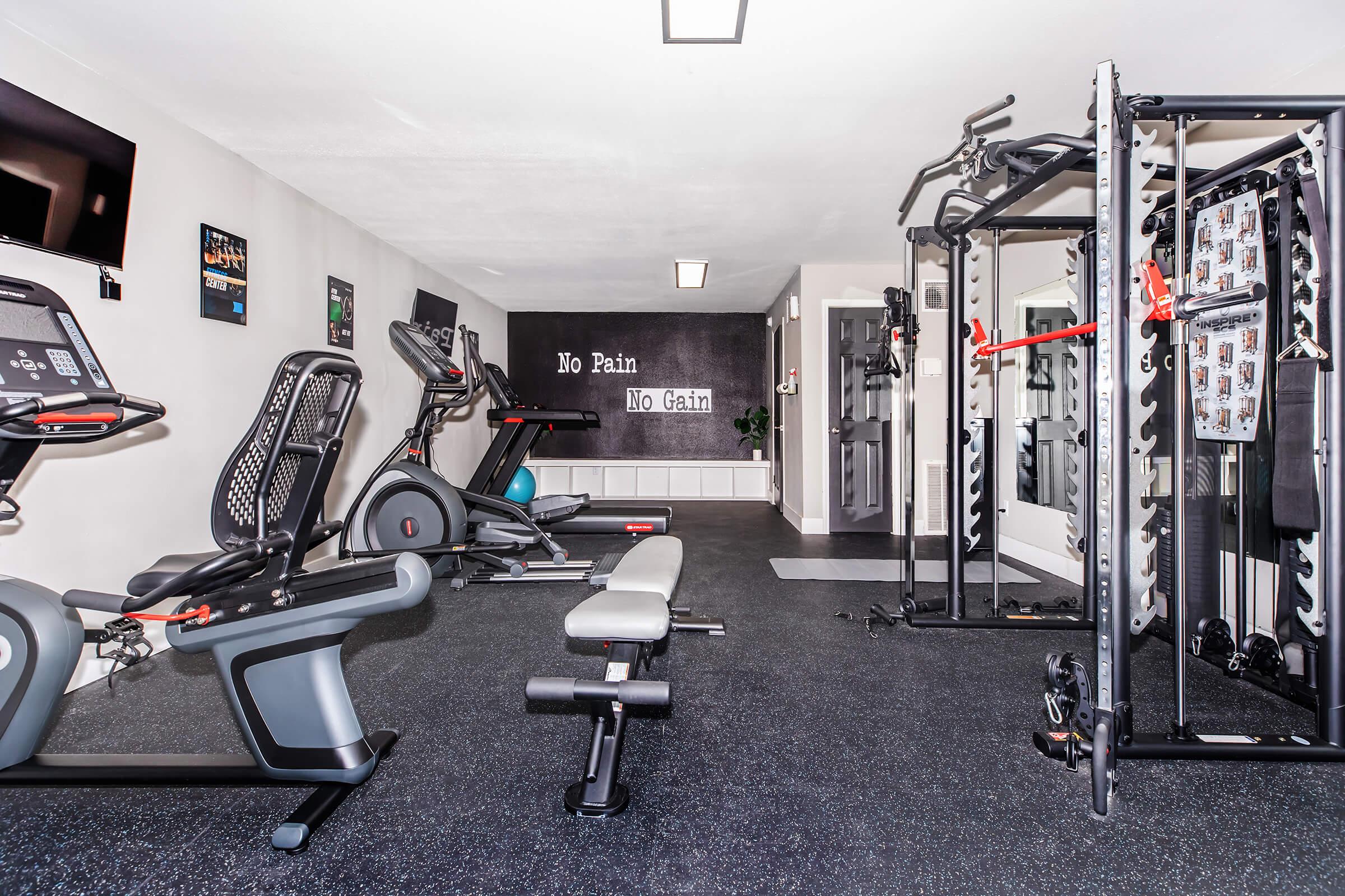 A well-equipped home gym featuring various exercise machines, including a treadmill, stationary bike, and multi-station weight rack. The room has black rubber flooring and motivational wall art that reads "No Pain, No Gain." Natural light comes from a window, creating a bright workout space.