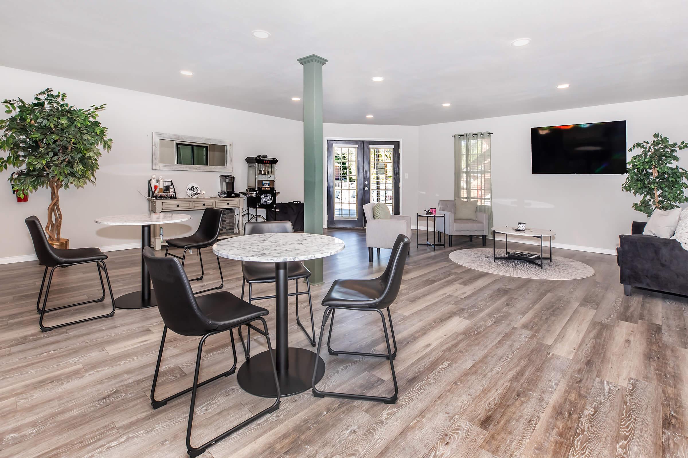 A modern, spacious living area featuring two high tables with stools, a cozy seating arrangement with a sofa, and a round coffee table. Large windows let in natural light, and there are potted plants adding greenery. A flat-screen TV is mounted on the wall. The flooring is light wood.