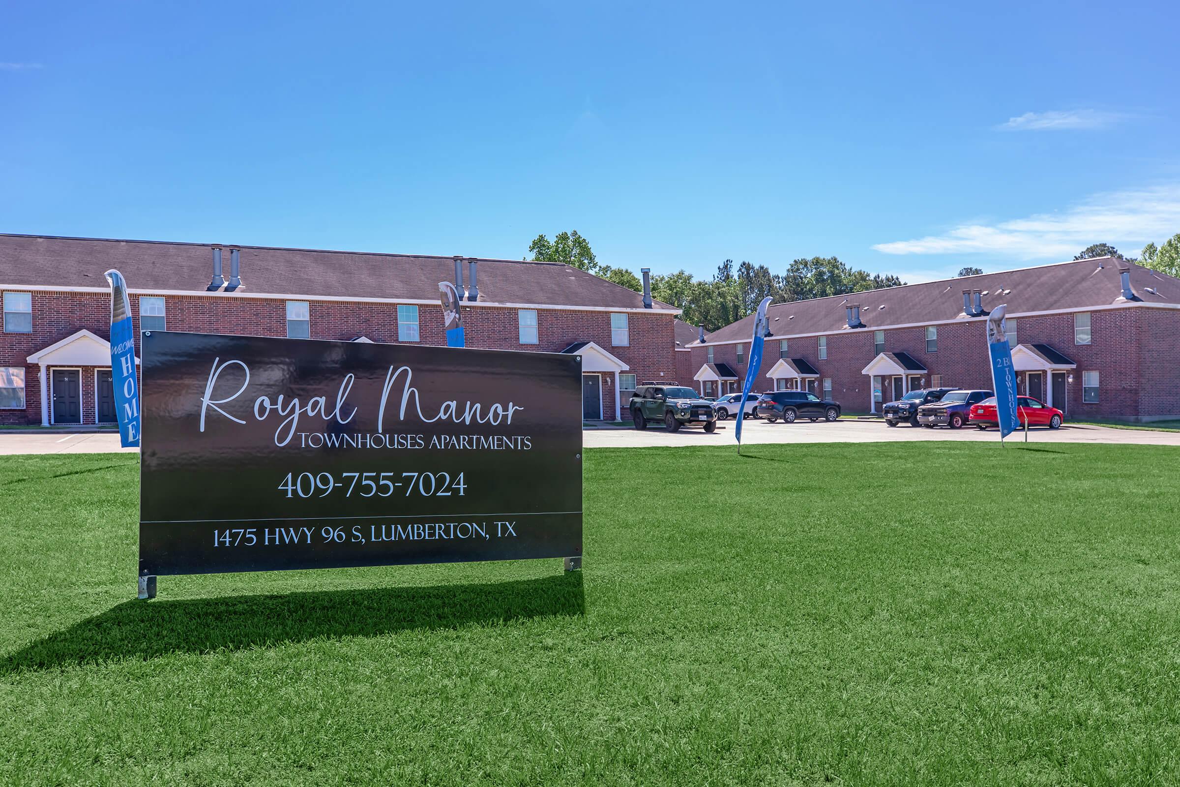 Sign for Royal Manor Townhouses Apartments located at 1475 Hwy 96 S, Lumberton, TX. The sign includes a phone number for inquiries. The background features several brick townhouse buildings and a well-maintained green lawn under a clear blue sky.