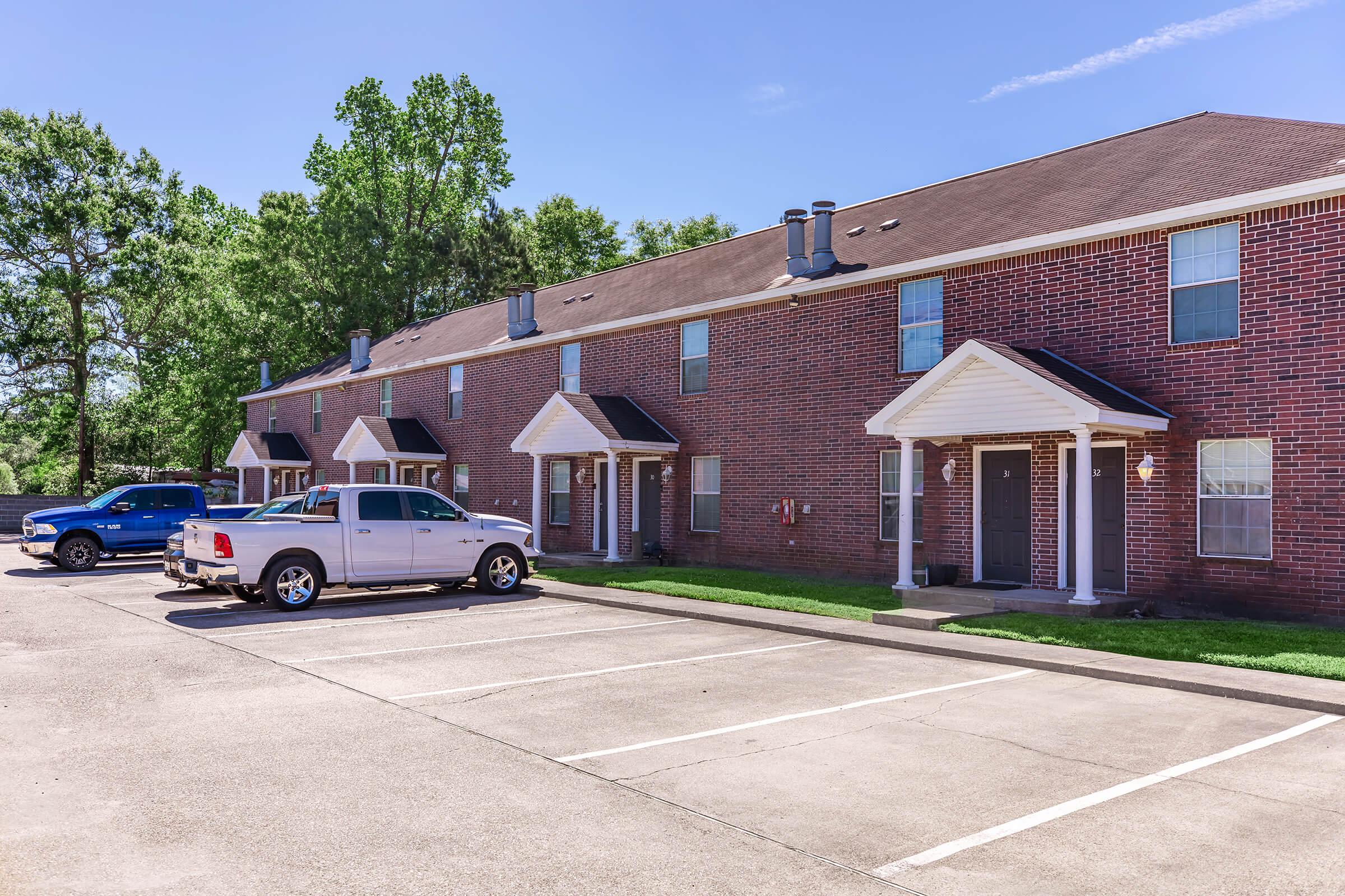 A row of brick apartment buildings with a well-maintained lawn in front. Several parking spaces are visible, occupied by a blue truck and a silver pickup. The sky is clear with a few clouds, and trees are visible in the background.