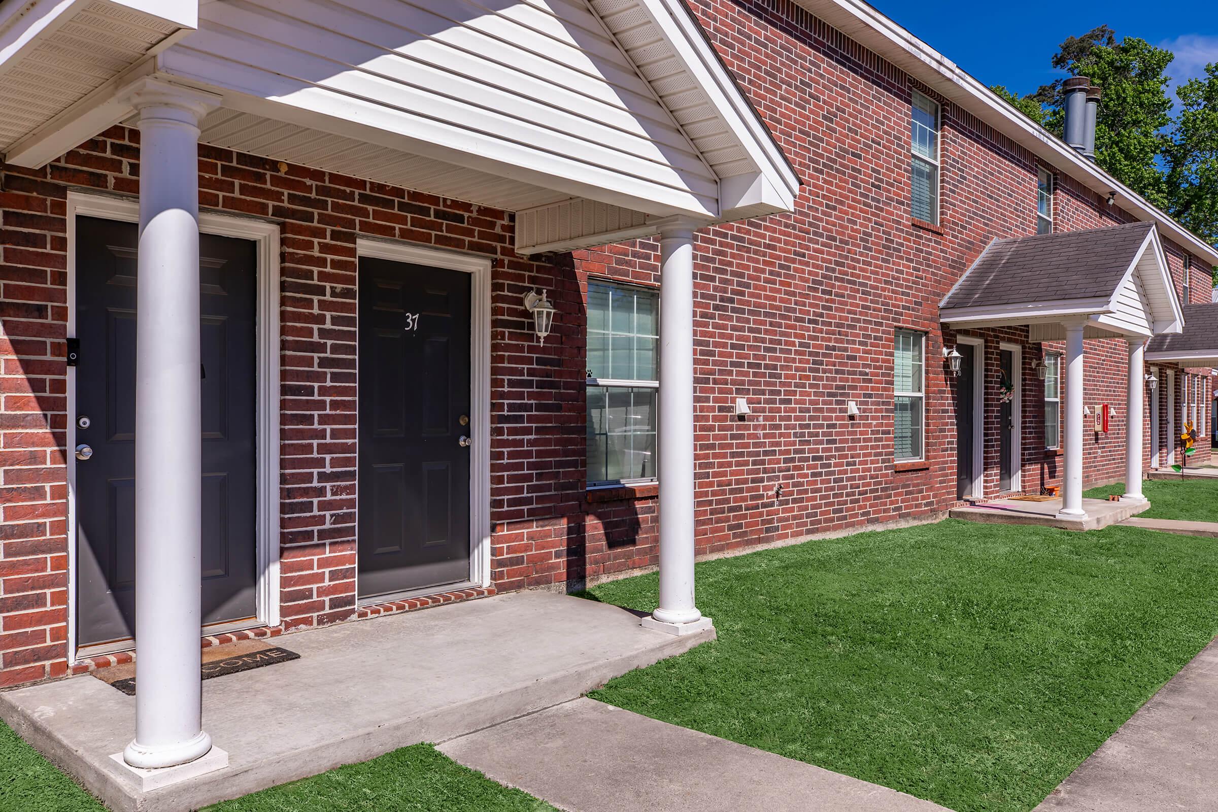 A row of brick townhouses featuring front porches, two main doors, and well-maintained green lawns. Each unit has a distinct door number, and outdoor fixtures like lamps are visible. The scene is bright and sunny, showcasing a well-kept residential area.
