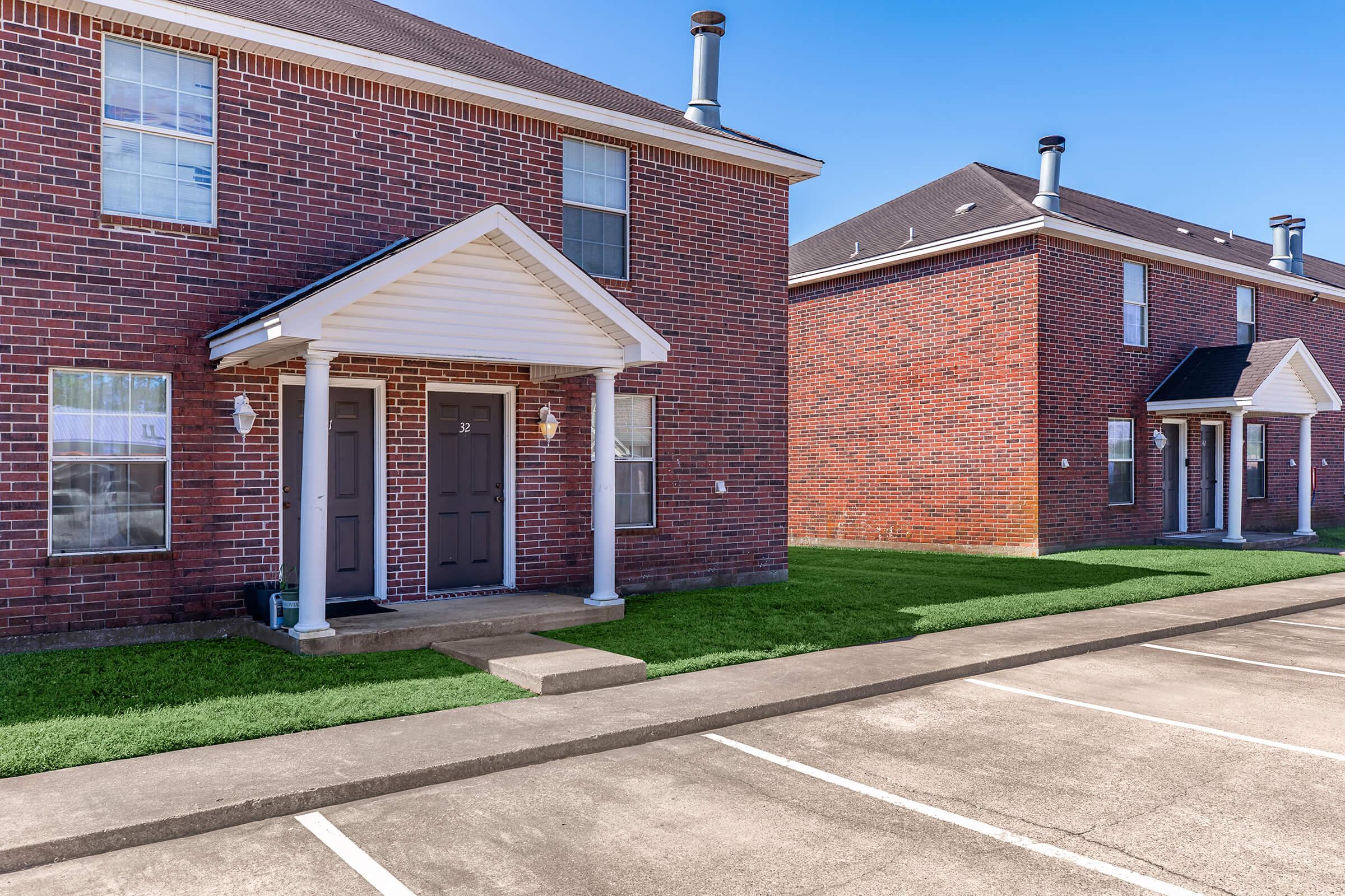Two brick apartment buildings with white trim and pitched roofs, featuring front entrances with small porches. Green grass lines the walkways, and a parking area is visible in the foreground. Clear blue sky above enhances the bright, sunny atmosphere.