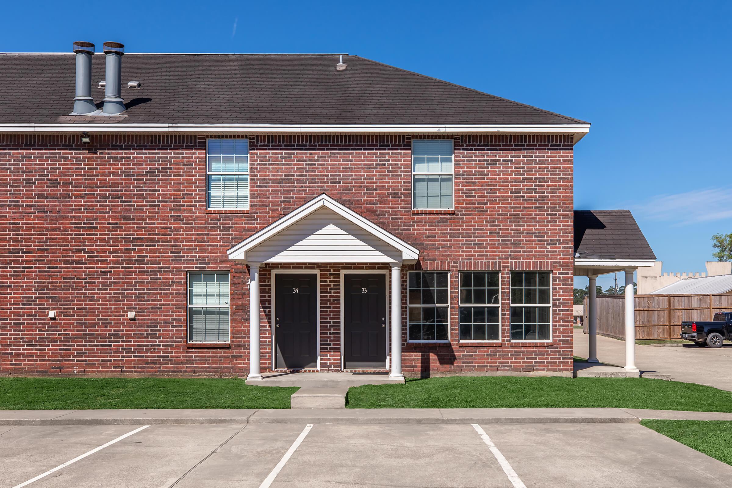 Two-story brick townhouse with a symmetrical facade, featuring two front doors and multiple windows. The property has a manicured lawn and is situated near parking spaces on a clear day.