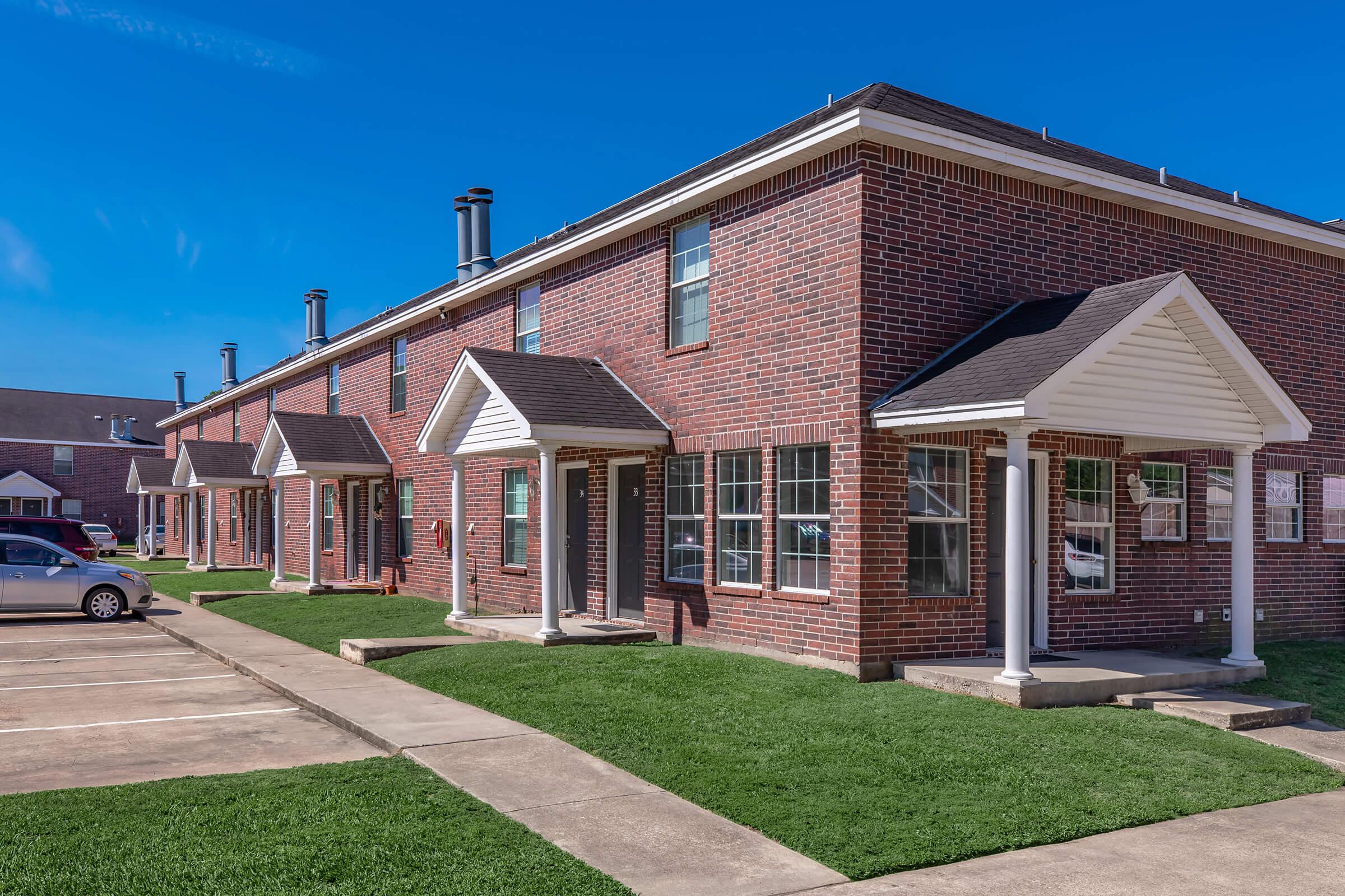 Brick apartment buildings under a clear blue sky, featuring multiple units with front porches and neatly trimmed grass. The scene includes a parking area with cars and concrete walkways leading to the doorways.