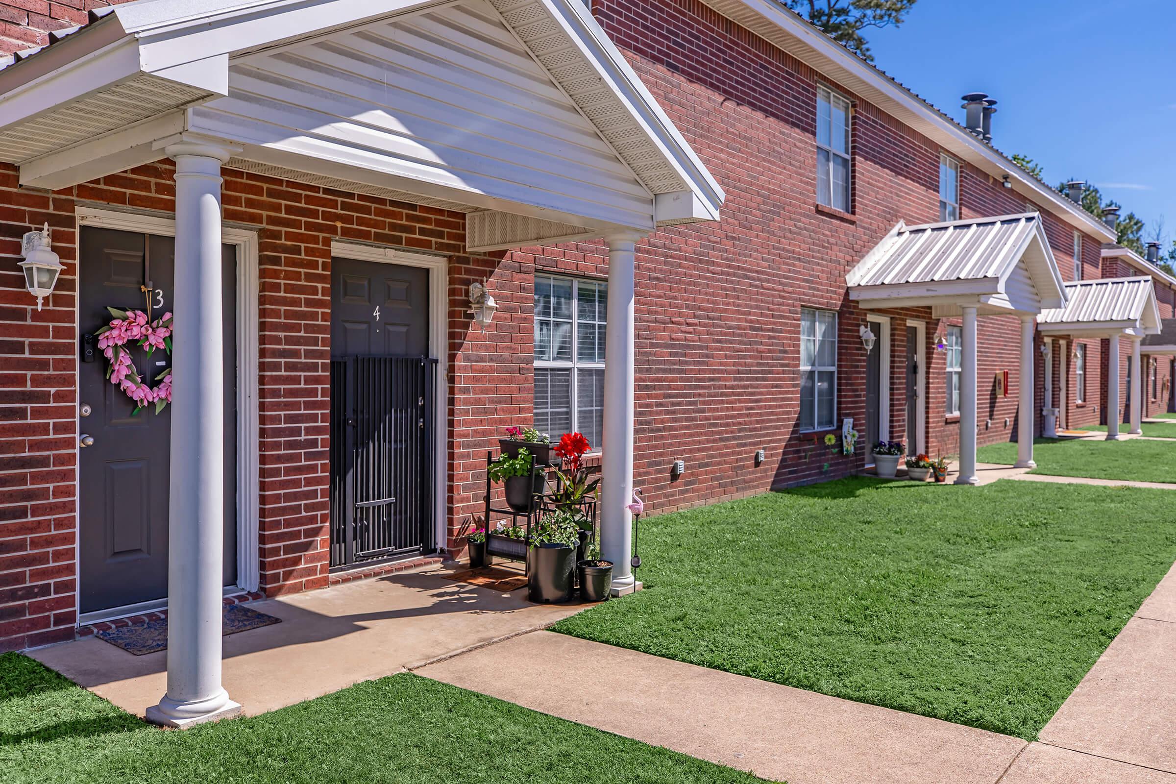 Row of brick apartment units with white awnings and wooden doors, each decorated with potted plants. Uniform green lawns separate the pathways, providing a tidy residential appearance on a sunny day.