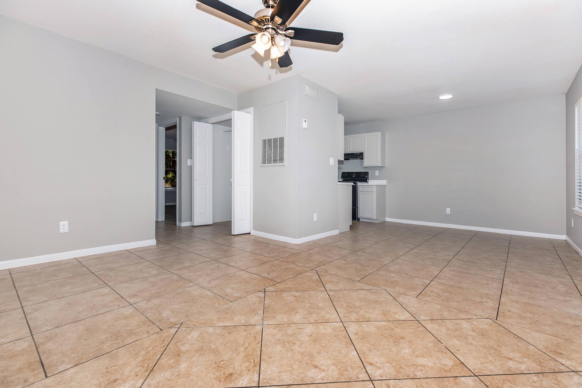 A spacious, modern living area with light gray walls and tiled flooring. A ceiling fan hangs overhead, and a kitchenette is visible in the background. The room features ample natural light and a clean, inviting atmosphere, making it ideal for relaxation or entertaining.