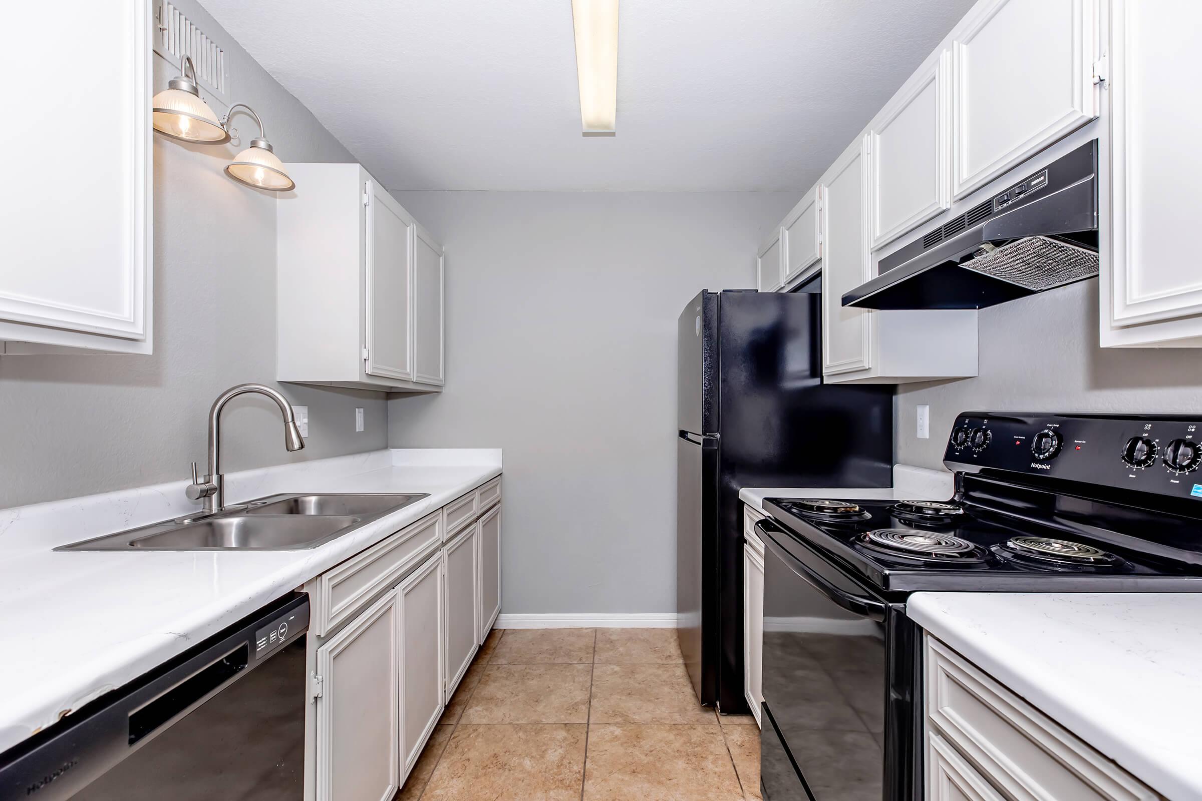 Modern kitchen featuring white cabinetry, stainless steel sink, and black appliances, including a refrigerator, oven, and stove. The countertops are light-colored, and the floor is tiled with beige tiles. The walls are painted a soft gray, and there are two overhead light fixtures.