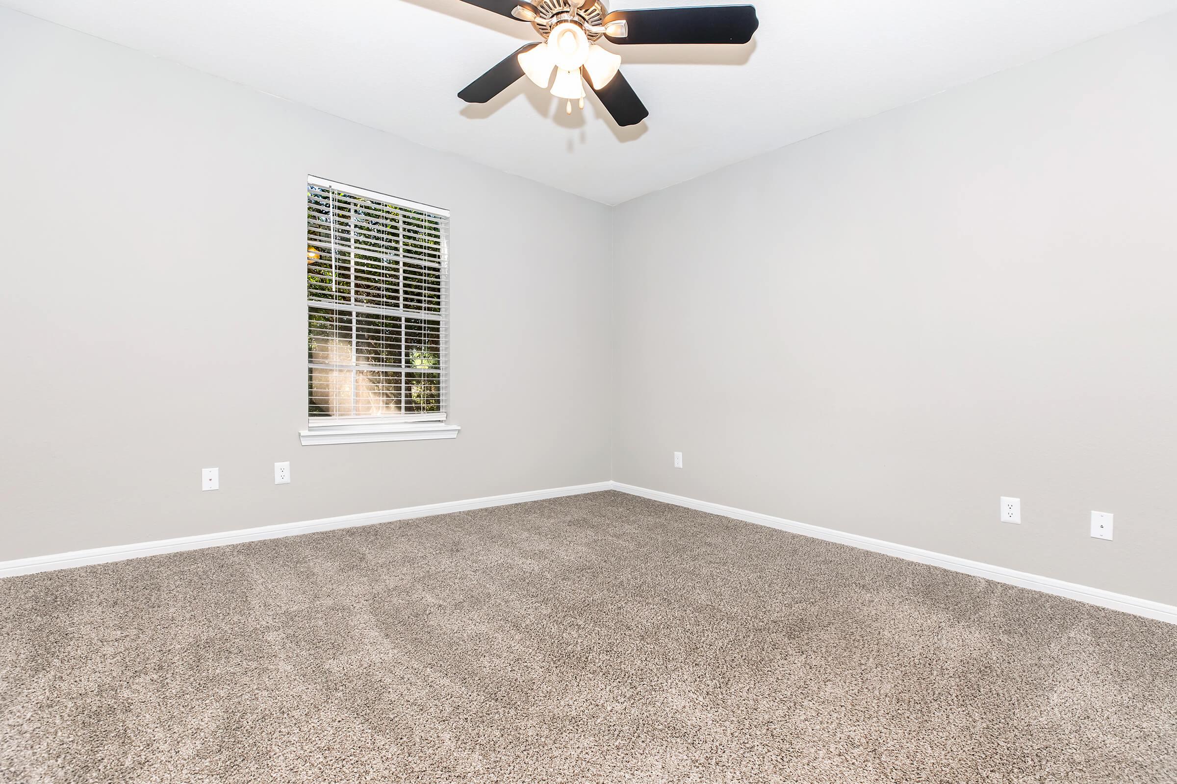 A spacious, empty room with light gray walls and plush beige carpet. There is a ceiling fan with four blades and a window with blinds allowing natural light to enter. The room features no furniture, creating an open and airy feel. The walls are undecorated, emphasizing the room's blank canvas for potential personalization.