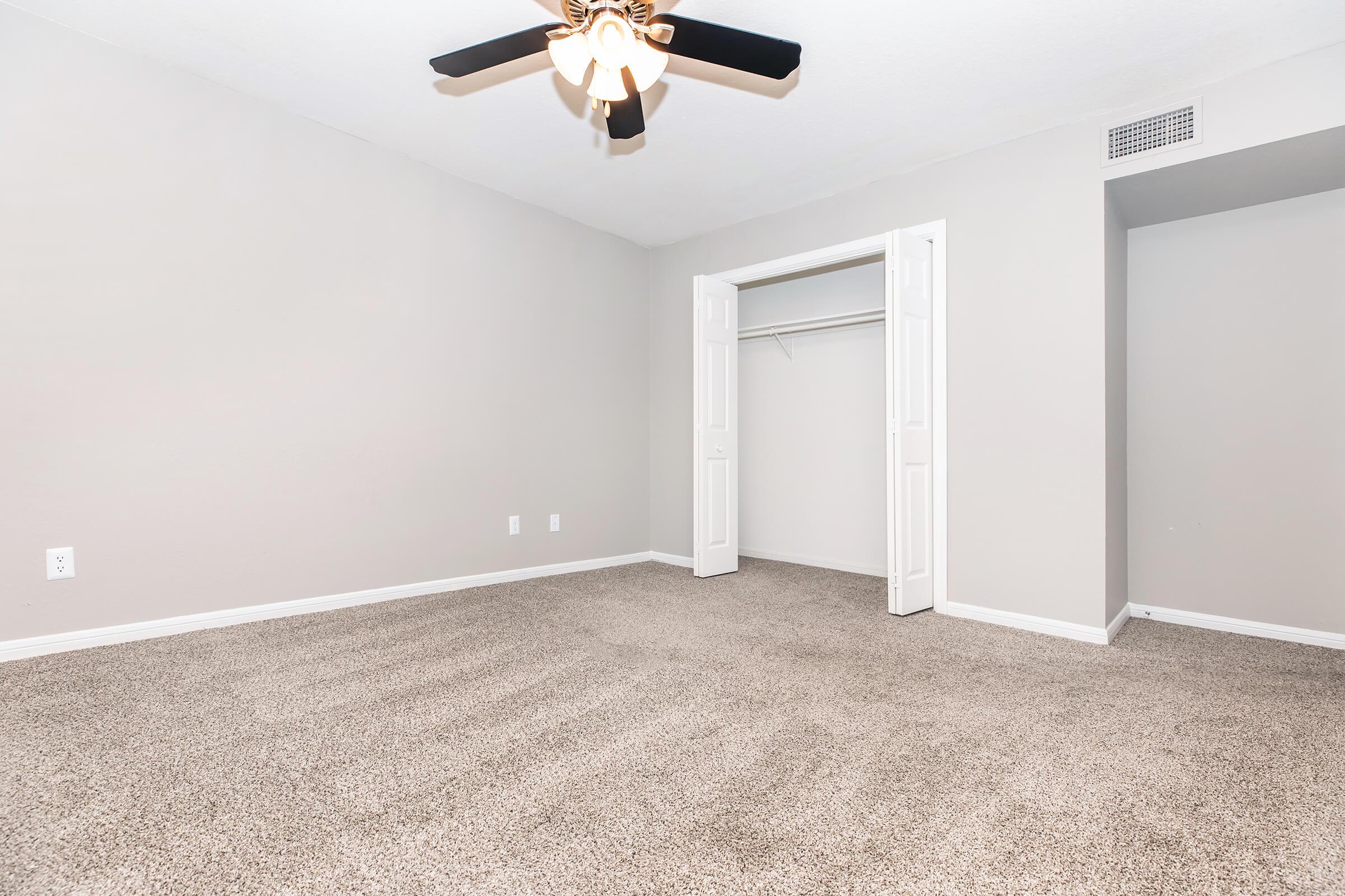 A spacious, empty bedroom with light gray walls and a light carpet. It features a ceiling fan with wooden blades and a closet with white double doors. Natural light from the window casts soft shadows, creating a clean and inviting space.