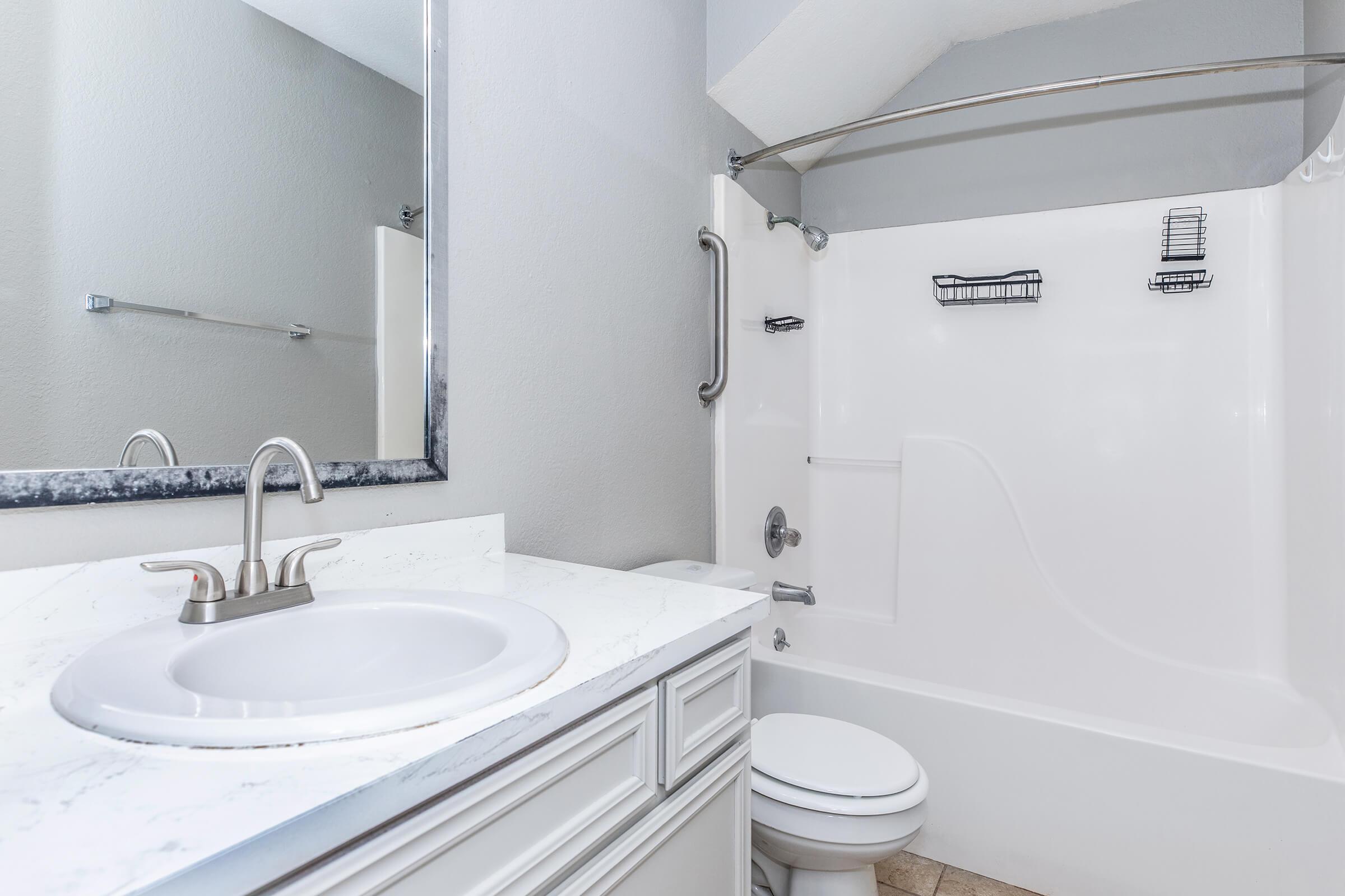 A clean bathroom featuring a white sink with a marble countertop, a mirror above the sink, a white bathtub with a shower, and grab bars. The walls are painted light gray, and there are shelves in the shower area for storage. The floor is tiled, creating a modern and functional space.