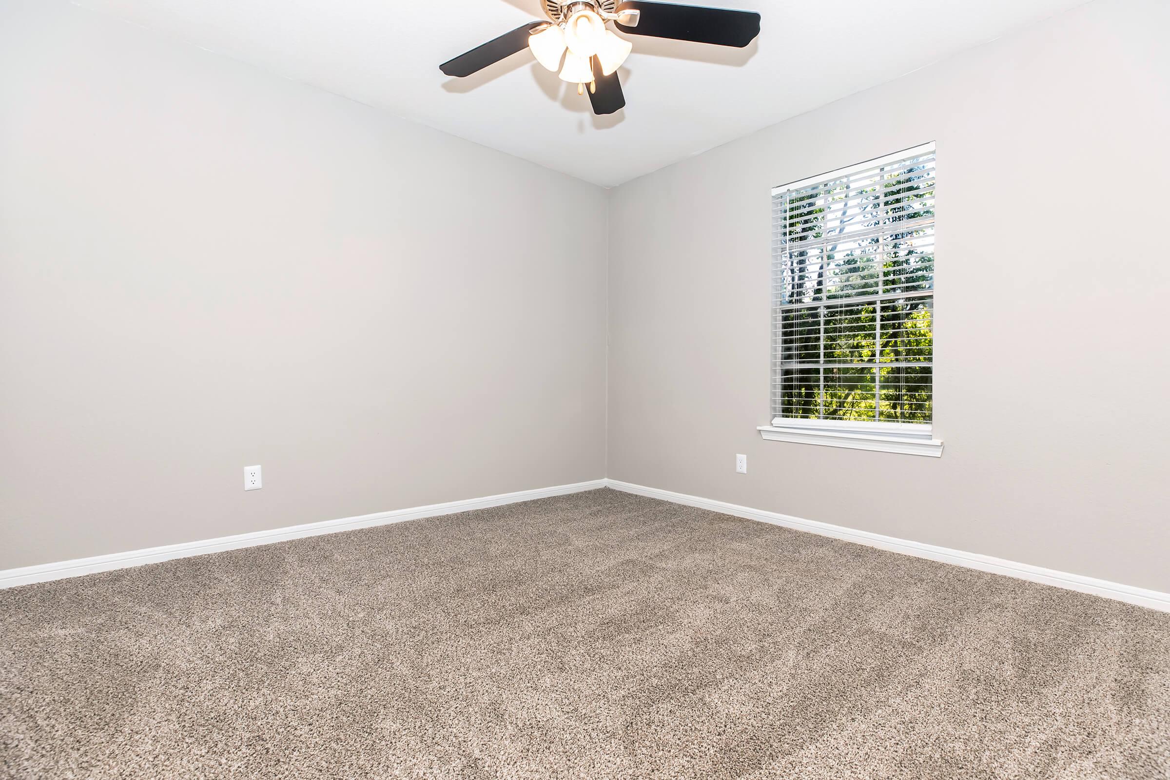Empty room with light gray walls, a ceiling fan, and a window with blinds. The carpet is a light beige color. Natural light is coming through the window, illuminating the space. The room has a simple and clean appearance, ideal for various uses.