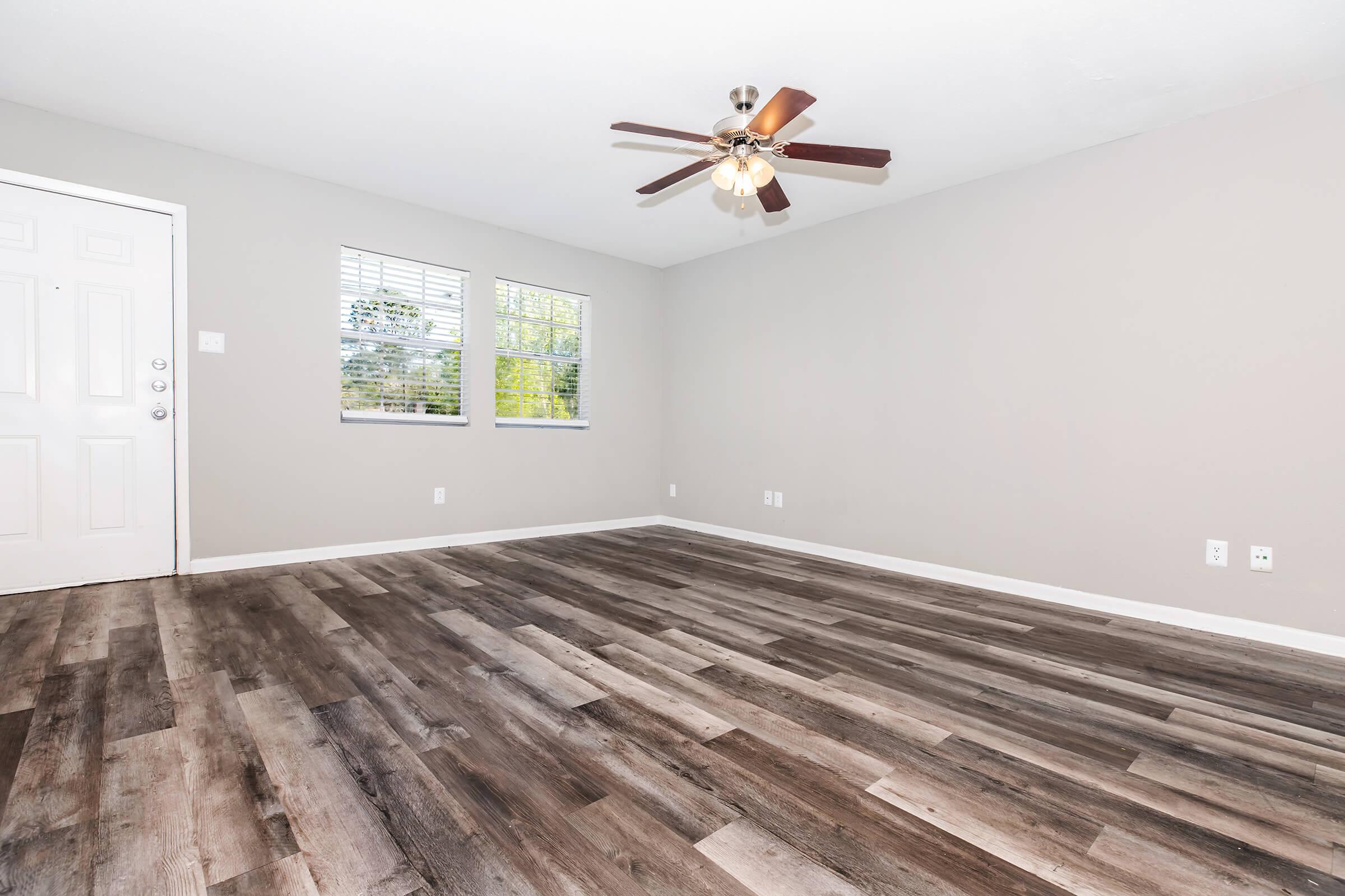Empty room with light gray walls and a ceiling fan featuring five lights. There are two windows letting in natural light, and the floor is covered in a wood-like laminate. A front door is visible on the left side, and there are no furnishings, creating a spacious and open atmosphere.