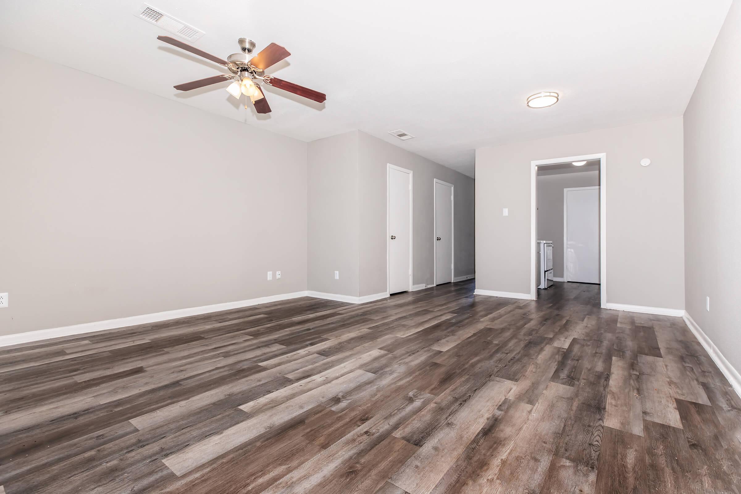 Spacious interior of a room with light-colored walls and a ceiling fan. The floor is covered in wood-looking laminate. There are doorways leading to other rooms, and the entrance to a bathroom is visible in the background. The overall ambiance is bright and welcoming.