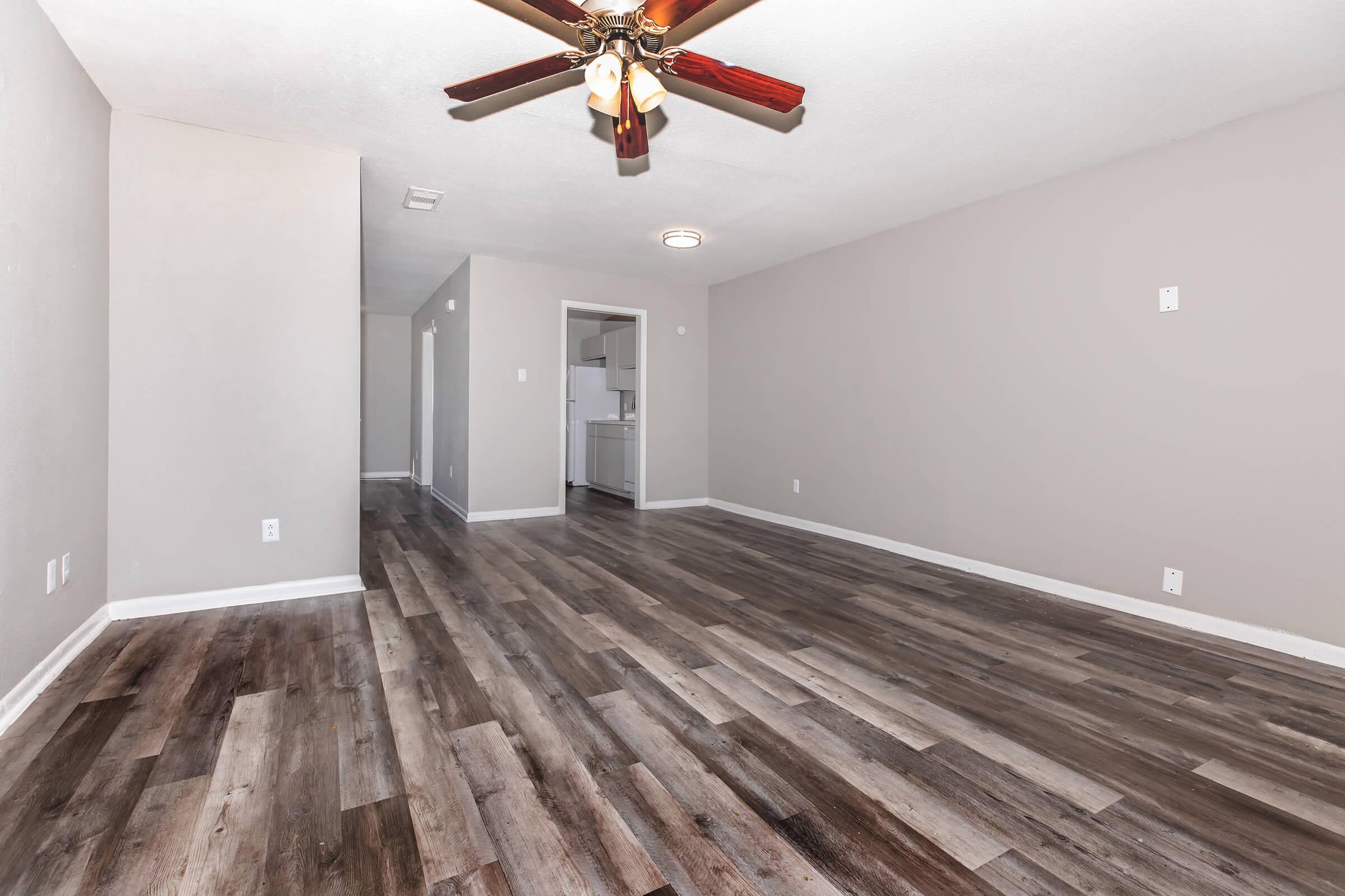 A spacious, empty living room with a ceiling fan and freshly painted light gray walls. The floor is covered in wood-like laminate. A doorway leads to a kitchen at the back, and there are other doorways on the left side of the image. Soft natural light illuminates the room, creating a bright atmosphere.