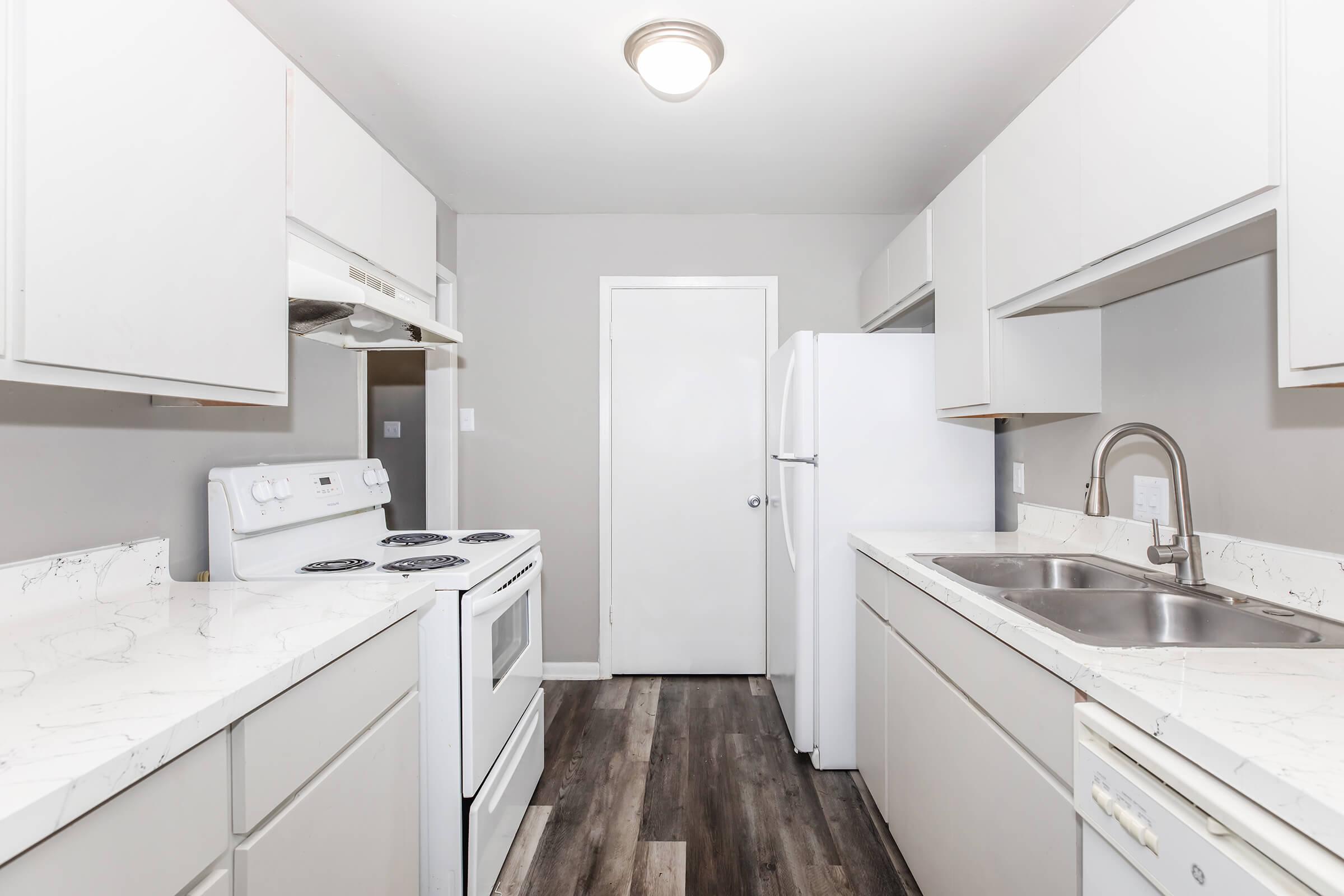 A modern kitchen featuring white cabinetry, a double sink, and stainless-steel appliances. The countertops are light-colored, with a subtle marble pattern. There is a stove, an oven, and a refrigerator, along with a door leading to another room. The flooring is dark wood, contrasting with the light walls and cabinets.