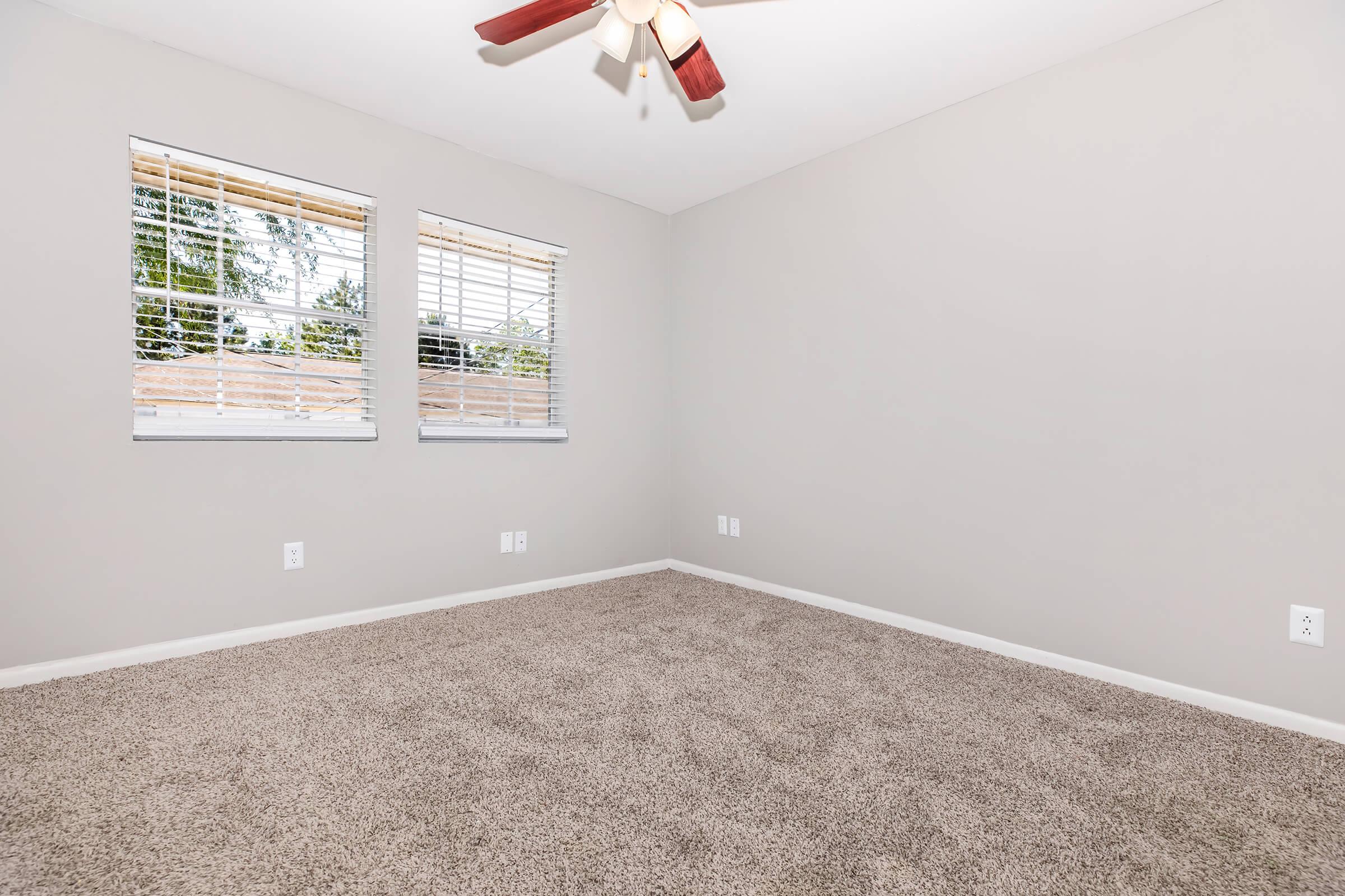Empty room with light gray walls and tan carpet. Two windows with blinds let in natural light. A ceiling fan with wooden blades is mounted in the center of the room. No furniture or decorations are present, creating a spacious and minimalistic atmosphere.