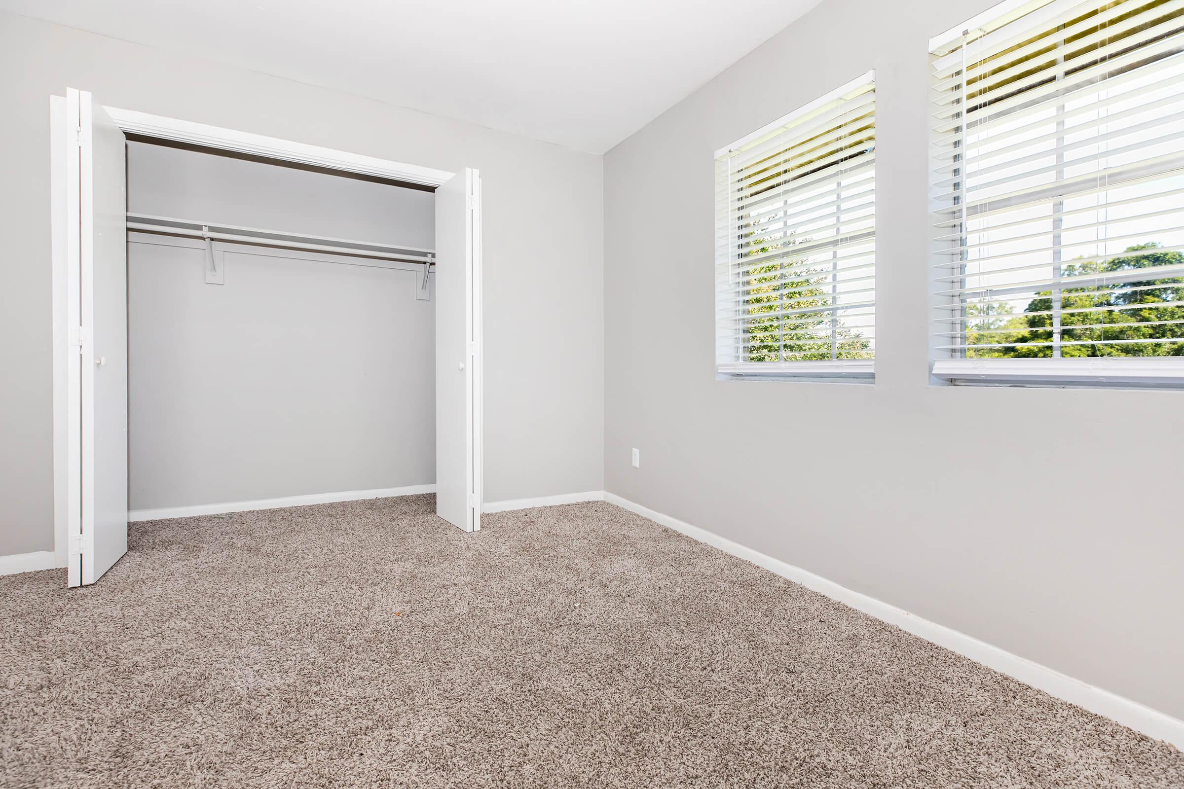 A bright, empty room with beige carpet and light gray walls. Two windows with white blinds allow natural light to fill the space. An open closet with a single hanging rod is visible on the left side. The room is uncluttered and has a clean, minimalist feel, ideal for a bedroom or office.