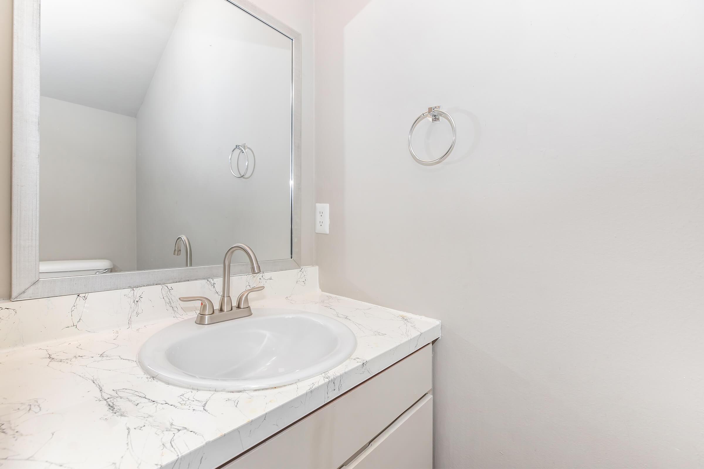 Bathroom sink area featuring a white countertop with marble pattern, a round sink, a modern faucet, and a mirror. A towel ring is mounted on the wall nearby, and the background is a plain light-colored wall. The overall ambiance is clean and minimalistic.