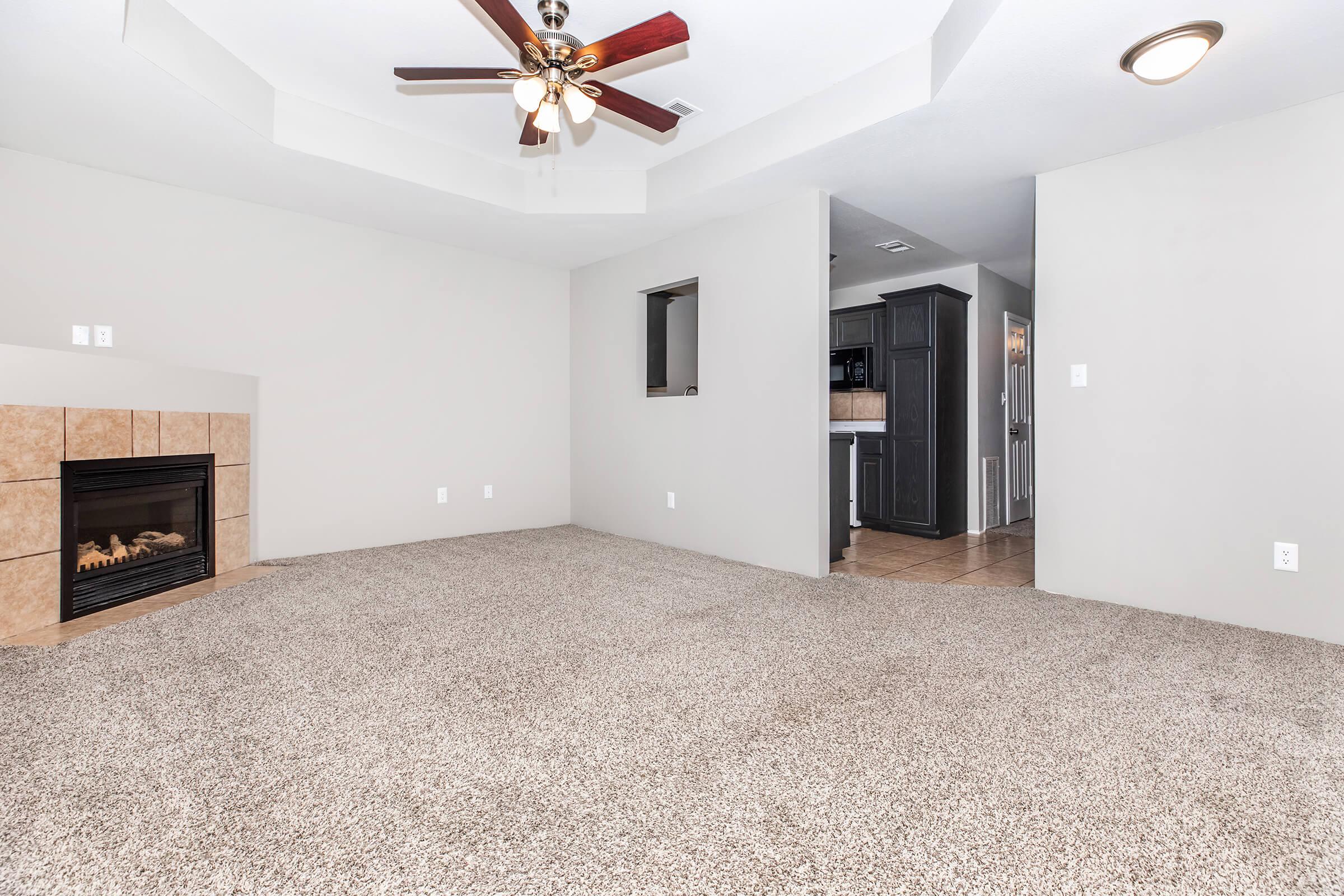 A spacious living room with plush carpet, featuring a ceiling fan and a gas fireplace on a tiled wall. A doorway leads to a kitchen area with dark cabinetry visible in the background. The walls are painted a light color, providing a bright and airy atmosphere.