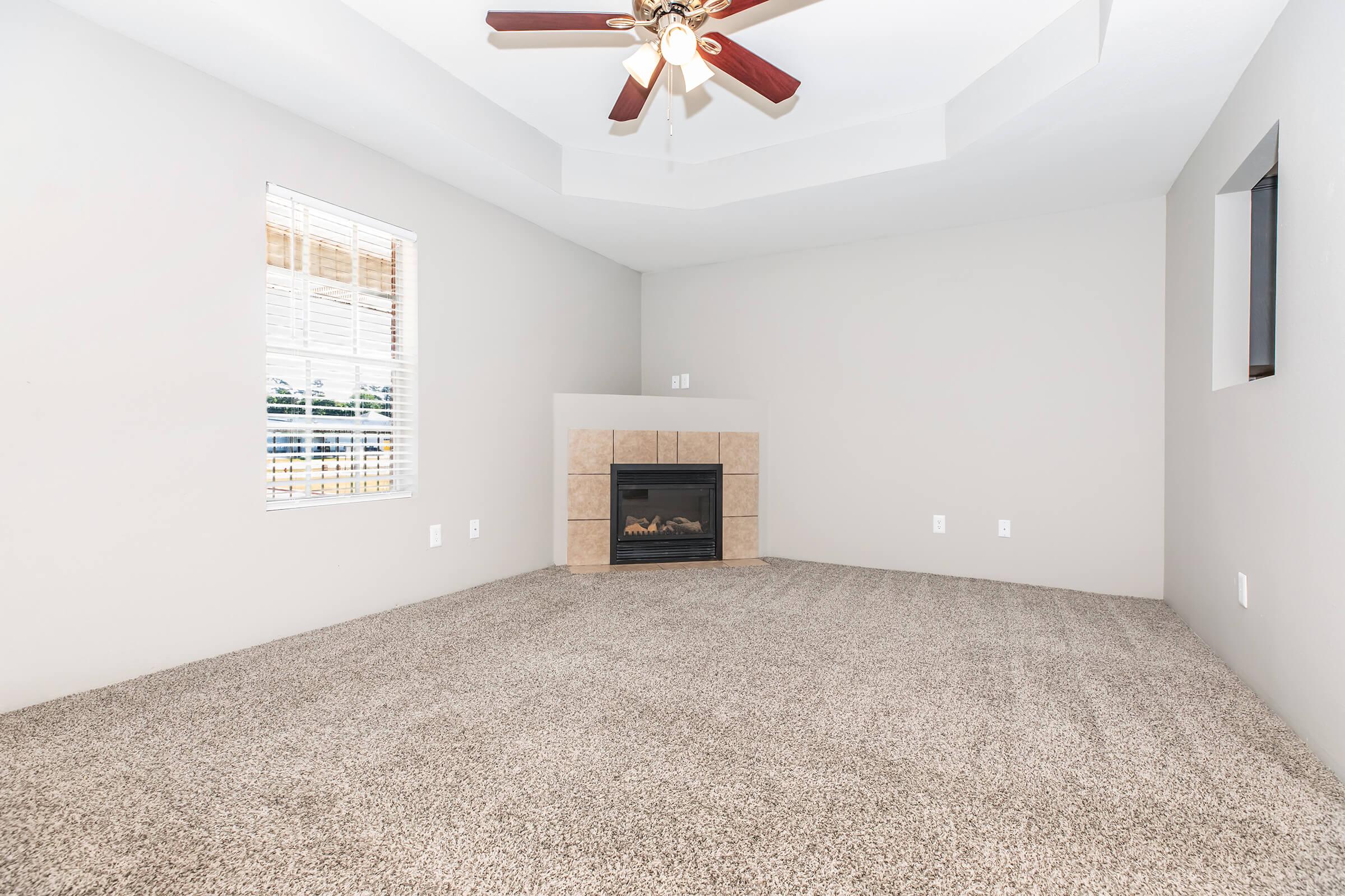 A spacious, empty living room featuring light gray walls and a carpeted floor. A ceiling fan with wooden blades hangs from a tray ceiling. A gas fireplace with a stone surround is positioned against one wall, and a large window allows natural light to enter, offering a view outside.
