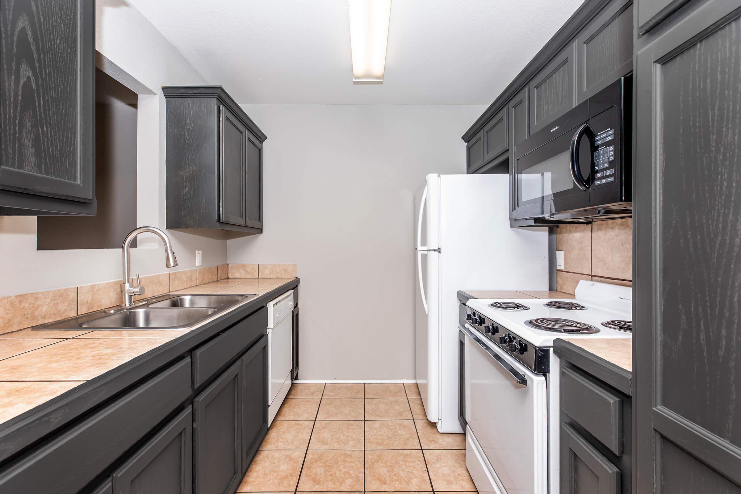 A modern kitchen featuring dark cabinetry, a double sink, a white refrigerator, an oven, and a microwave. The countertops are tiled, and the floor is covered with square tiles. Natural light from a ceiling fixture illuminates the space.
