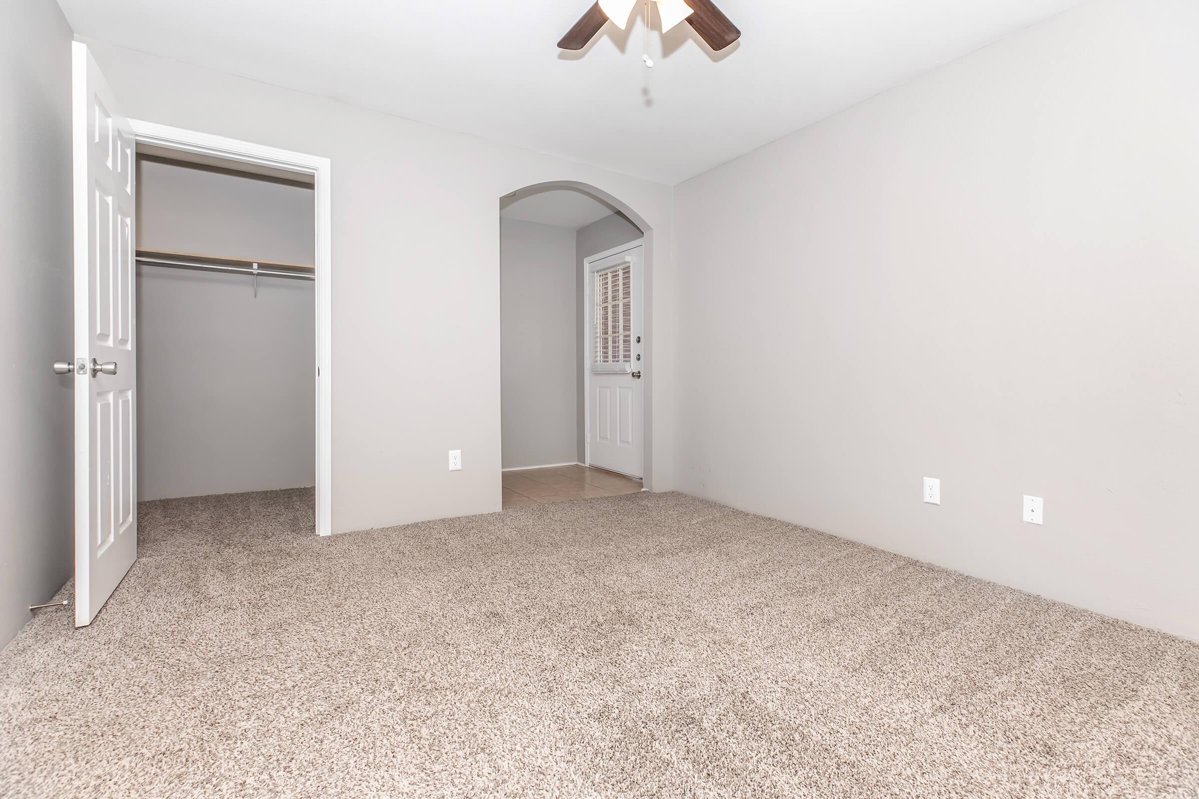 A spacious room with light gray walls and plush carpet. It features a ceiling fan and an archway leading to a small hallway with a door. There is an open closet on one side, providing additional storage space. The overall atmosphere is bright and inviting.