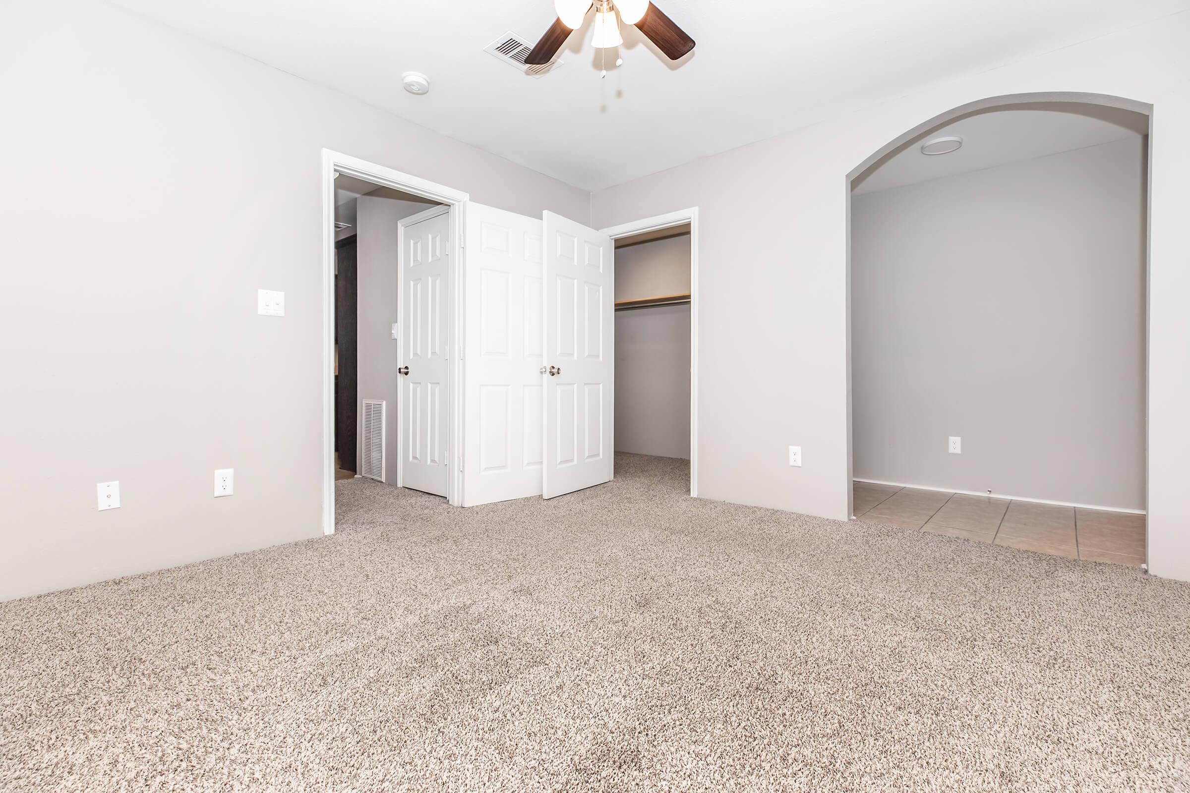 A spacious room with light gray walls and plush carpet. There are two doorways leading to adjacent spaces, one of which has a closet with folding doors. A ceiling fan hangs above, and the floor is mostly bare with a tiled section visible. The overall ambiance is bright and open.