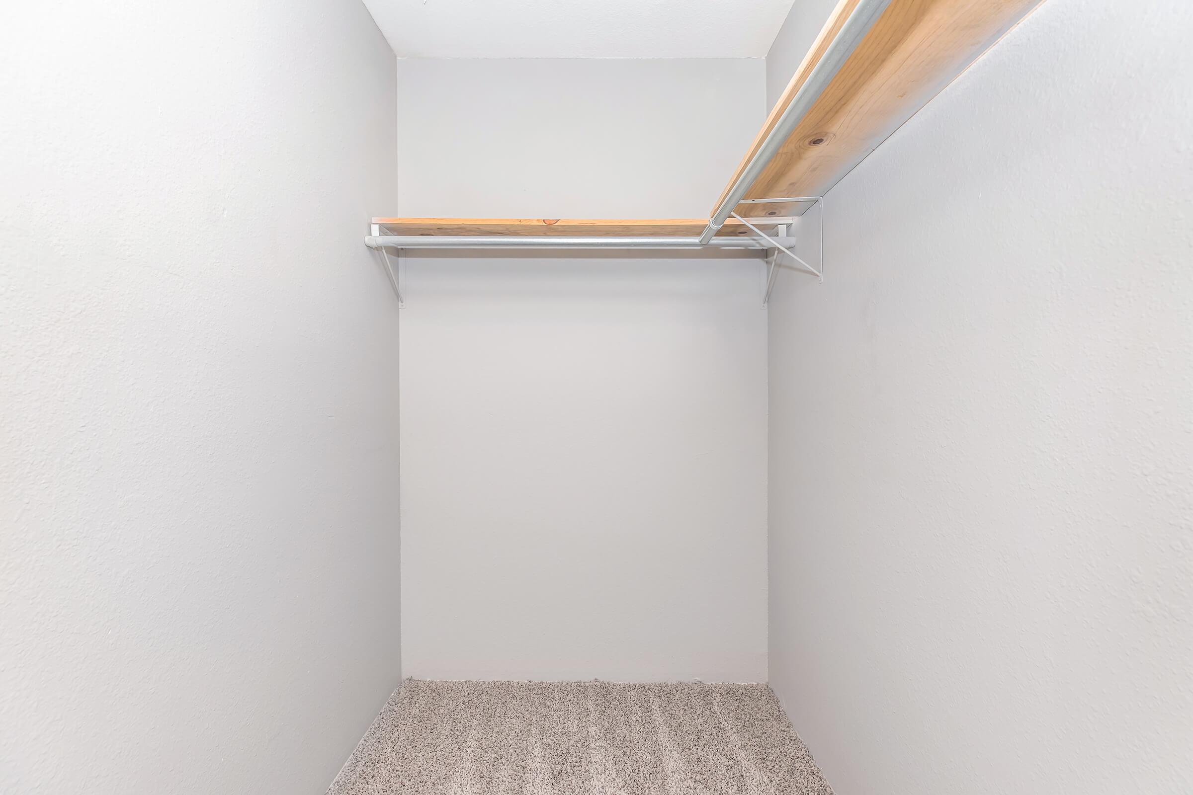 Empty closet with a white wall and a wooden shelf supported by a metal frame. The floor is carpeted in a light tone, enhancing the spacious appearance of the closet.