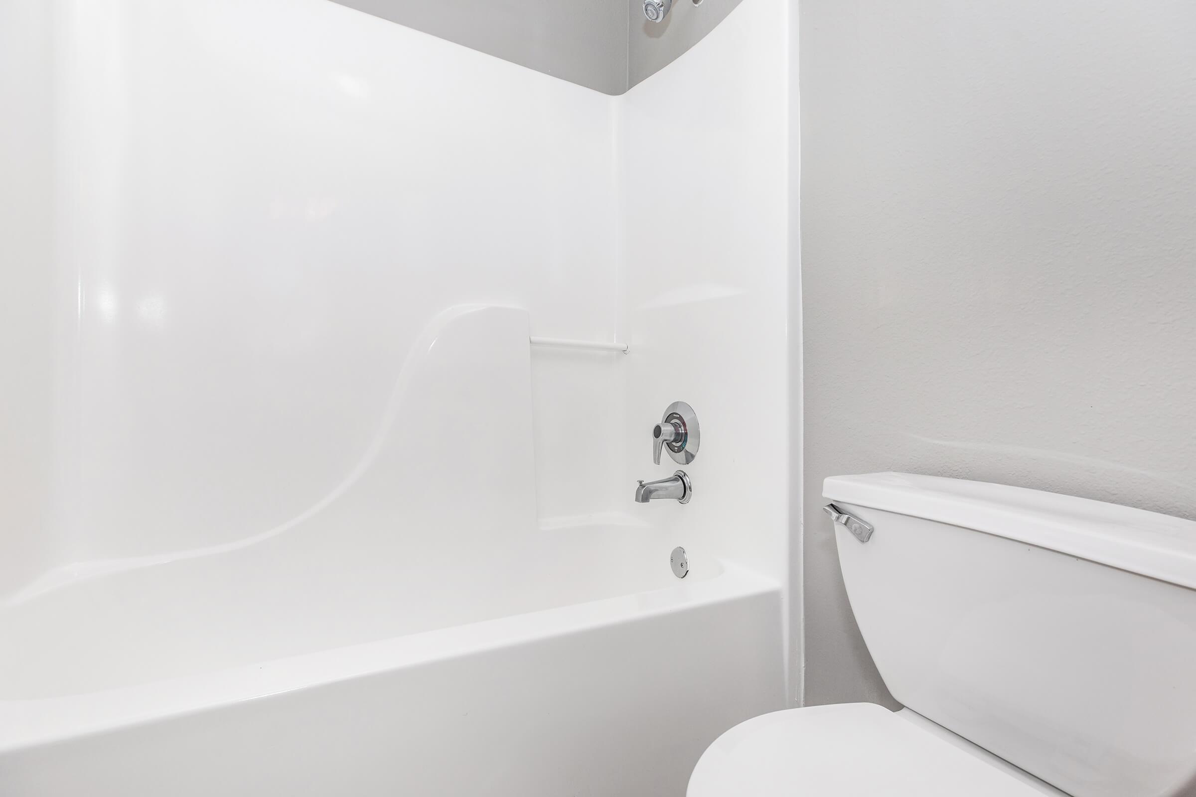 A clean and modern bathroom featuring a white bathtub with a smooth, curved design and a wall-mounted faucet. Next to it is a white toilet with a simple, contemporary appearance. The walls are painted in a light gray color, creating a bright and airy atmosphere.