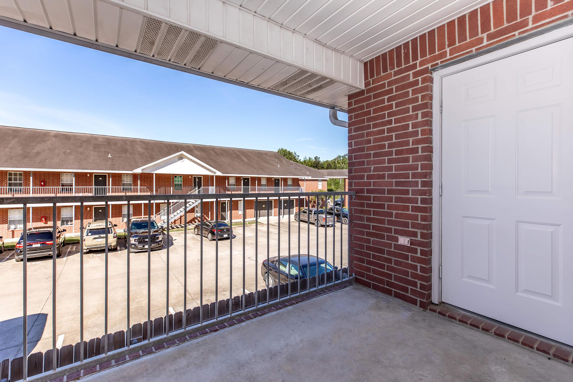 View from a balcony showing a multi-level apartment complex with a parking lot below. The scene includes several parked cars and a clear blue sky, framed by a simple brick railing and wall. The white door visible is typical of apartment entryways.