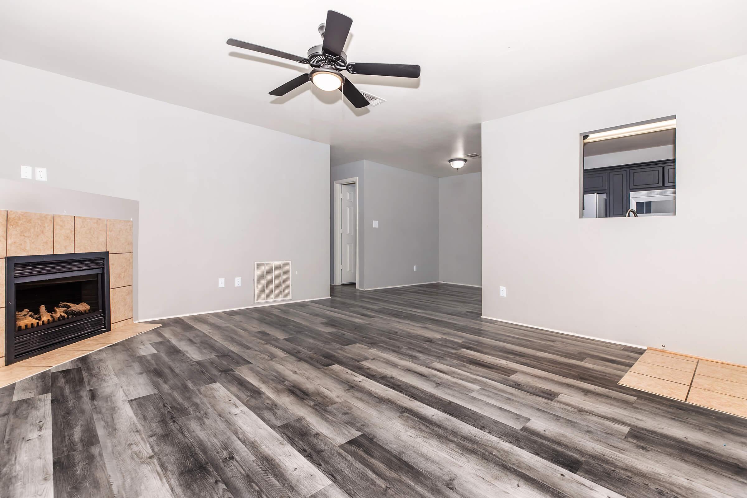 A spacious, empty living room featuring gray walls, a ceiling fan, and a modern fireplace. The floor is covered in wood-like laminate planks, and there's a small opening leading to another room in the background. Natural light comes from the window area, creating a bright and inviting atmosphere.