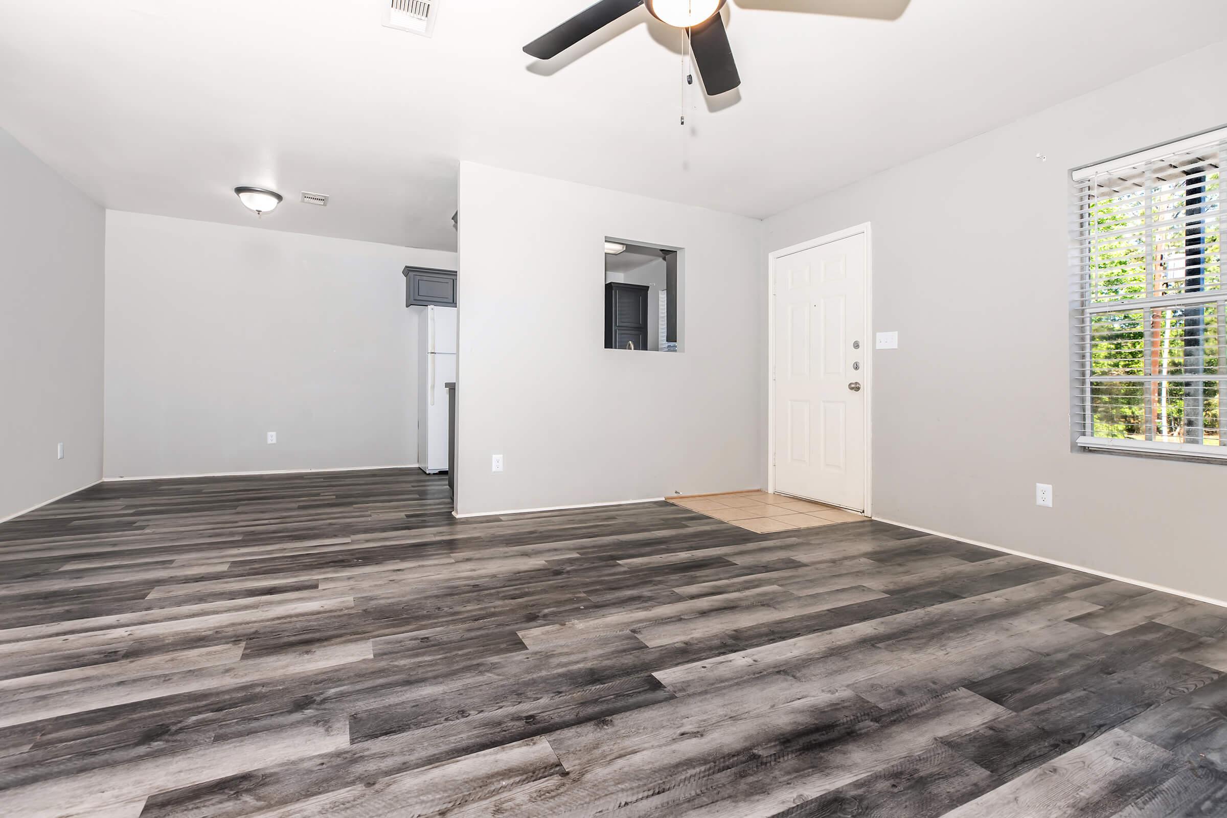Spacious room with gray walls, new wood-style flooring, and a ceiling fan. Features a doorway leading to an entrance, a window with blinds allowing natural light, and a small opening to an adjoining kitchen area.
