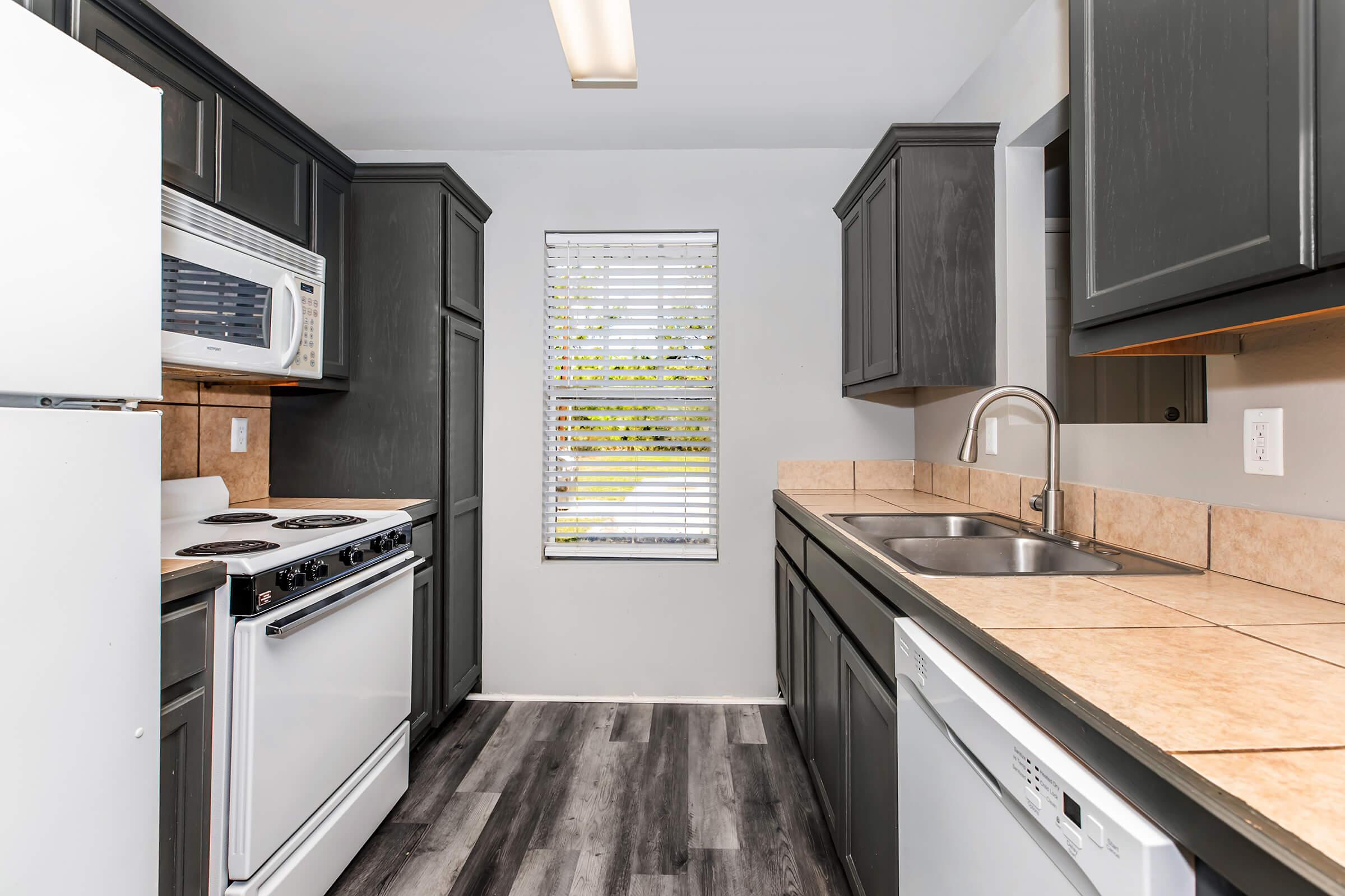 Modern kitchen featuring dark cabinetry, a white refrigerator, a built-in microwave, and a stove. There are two sinks with a chrome faucet and beige tile countertops. A window with blinds allows natural light, and the flooring has a wood-like appearance.
