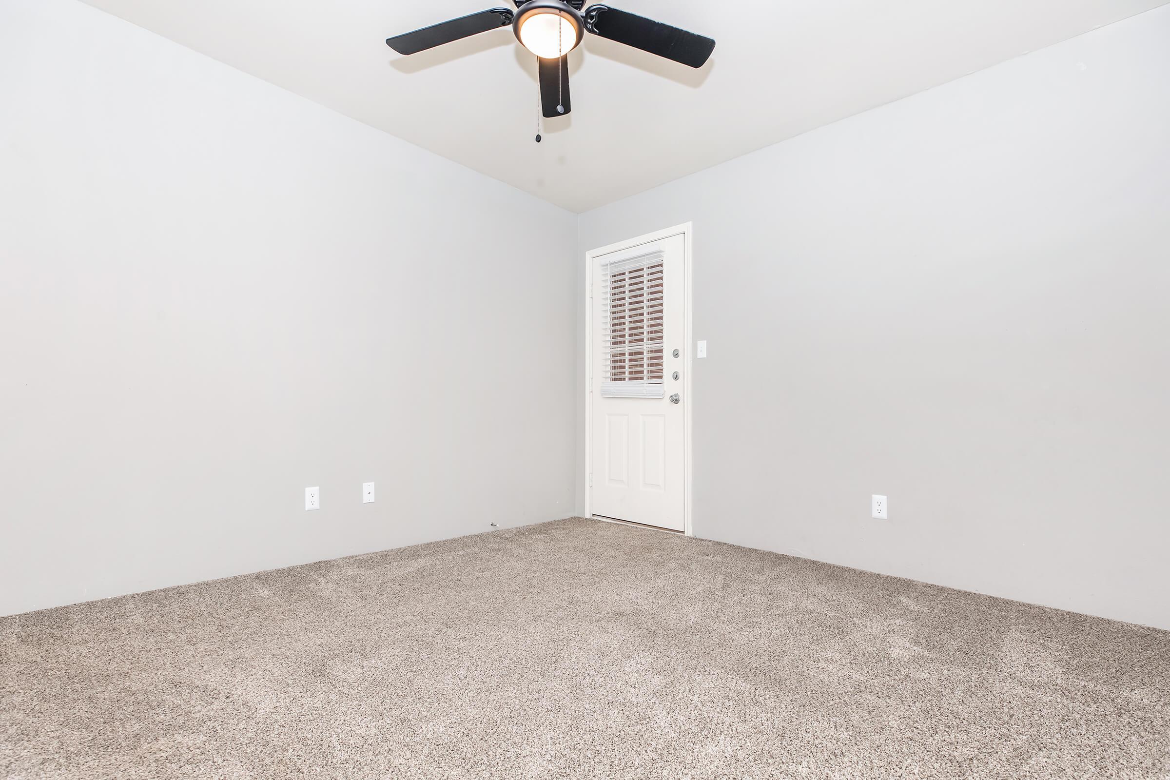 Empty room with light gray walls and beige carpet, featuring a ceiling fan. A door with a window is on the right side, and there are two electrical outlets on the wall. The space is well-lit and devoid of furniture or decor.
