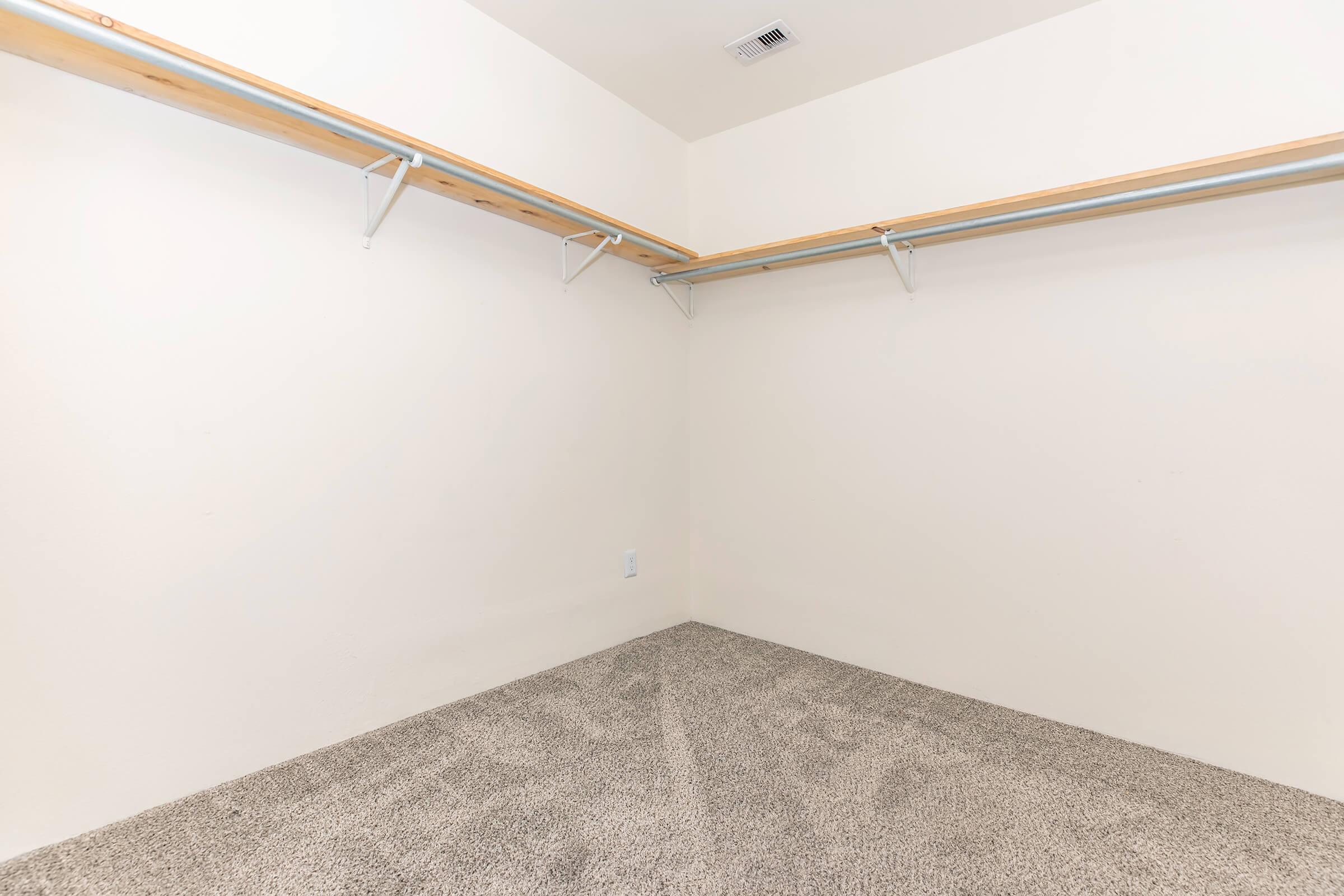 Empty closet with two wooden shelves mounted on metal brackets. The walls are plain white, and the floor is covered with beige carpet. There are no items or furnishings visible, creating a spacious and minimalist appearance.