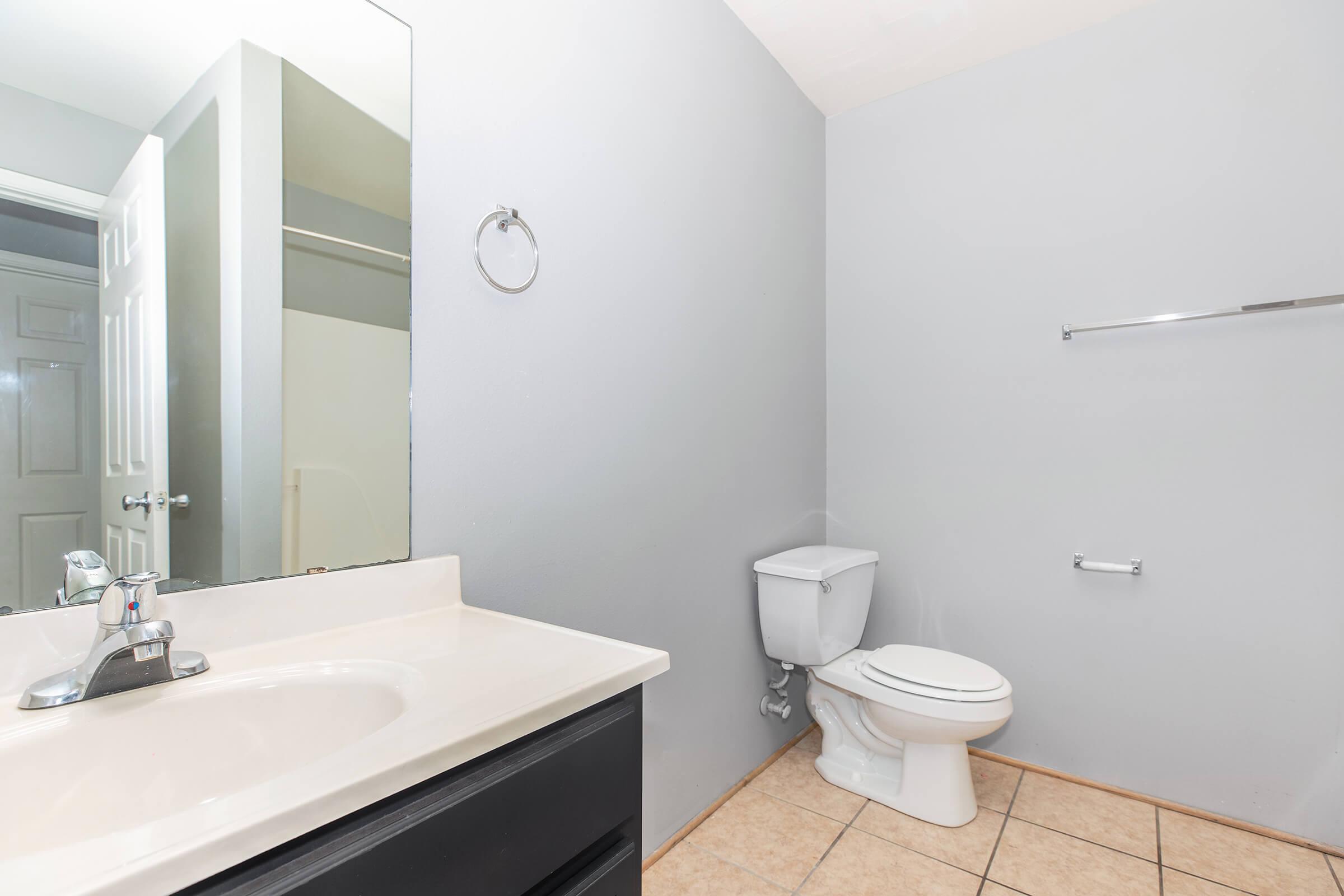 A clean and simple bathroom featuring a white sink with a mirror above, a toilet, and tiled flooring. The walls are painted light gray, and a towel ring is mounted near the sink. A door leads to a separate area, and a silver grab bar is present on the wall next to the toilet.