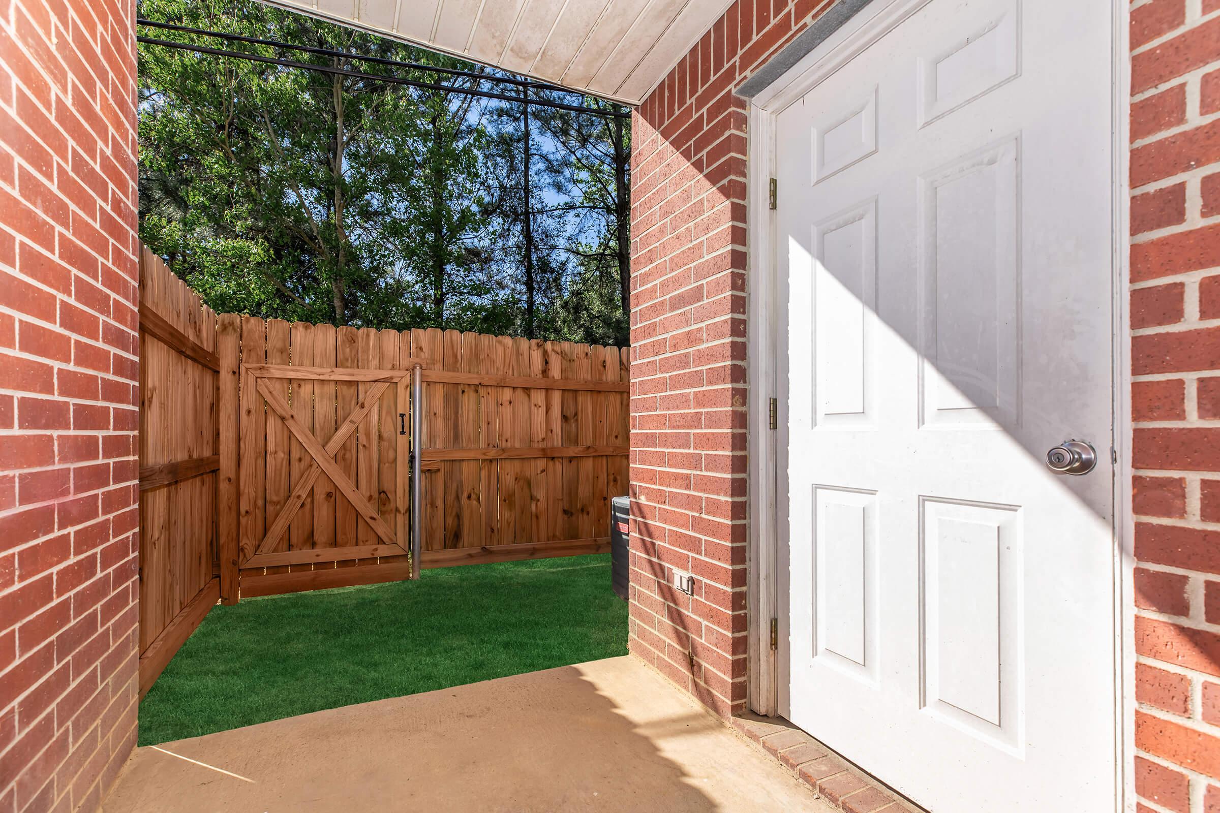 View from a covered entryway showing a white door and a fenced backyard. The walls are red brick, and there is a grassy area visible through a wooden gate in the fence. Natural light casts shadows on the ground, creating a warm and inviting atmosphere.