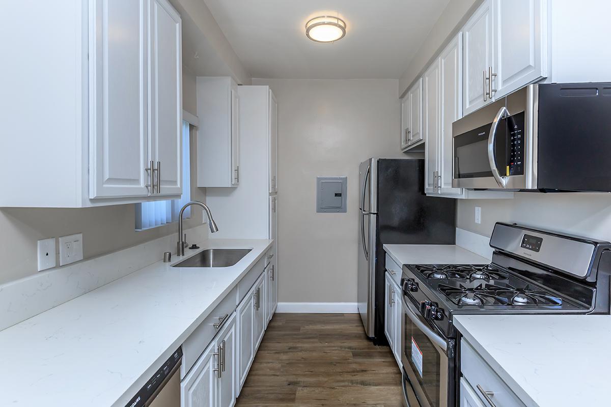Galley kitchen featuring goose neck faucet, white countertops and cabinets nested around stainless appliances. the gas stove and overhead microwave is showcased here.