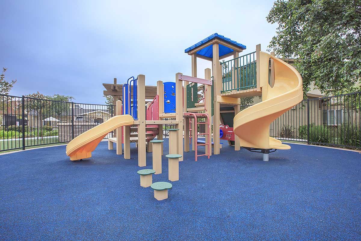 A colorful playground structure featuring slides, climbing areas, and various play panels, set on a blue rubber surface surrounded by a safety fence and green grass.