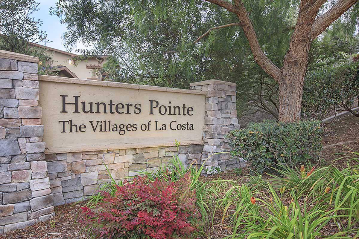 Sign for "Hunters Pointe" at "The Villages of La Costa," featuring natural stone elements and surrounding greenery, including shrubs and flowers.