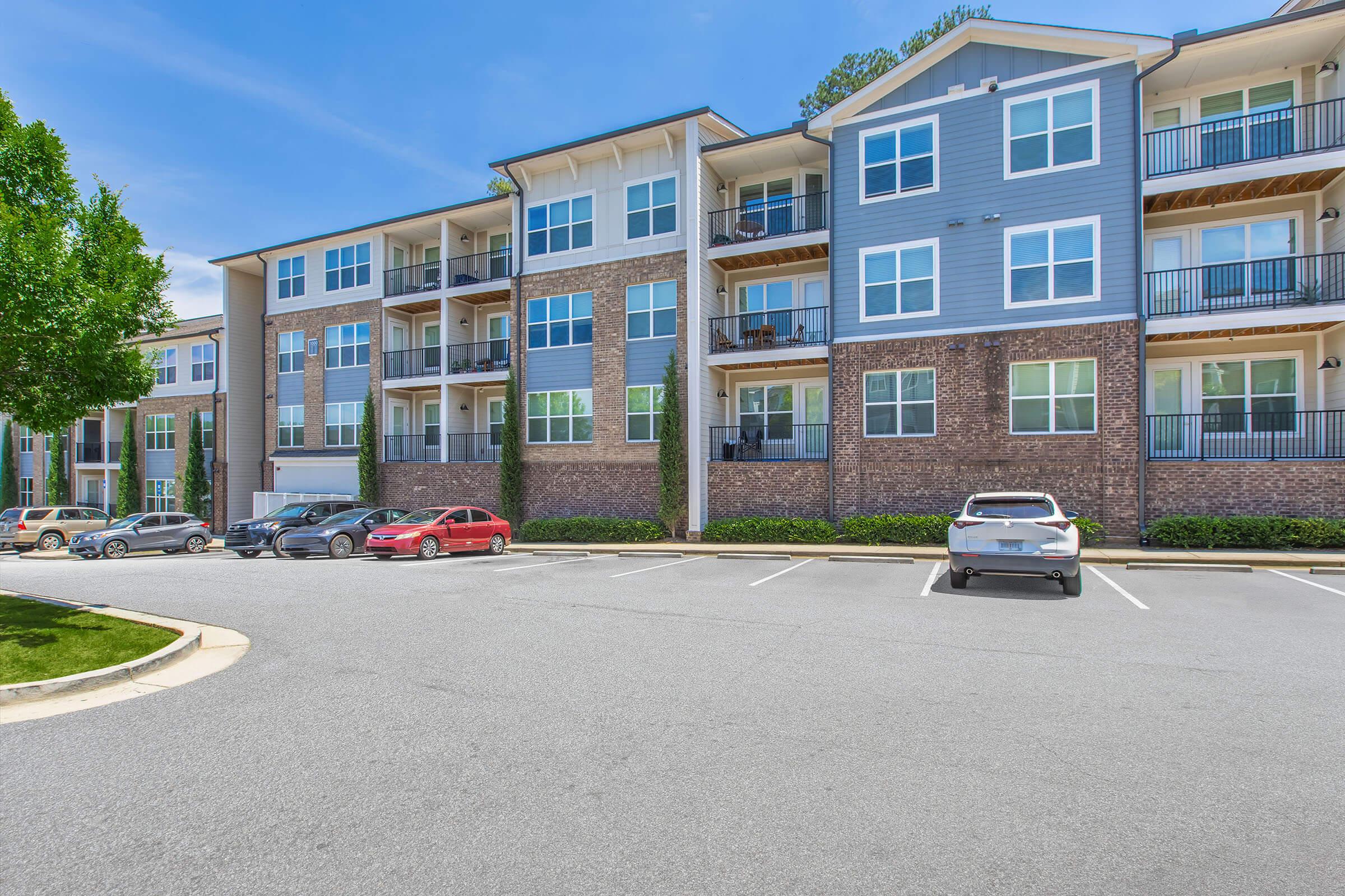 A modern apartment building with multiple stories, featuring a mix of brick and blue siding. Balconies are visible on each level, and there are several parked cars in the foreground. The setting includes well-maintained landscaping and a clear blue sky.