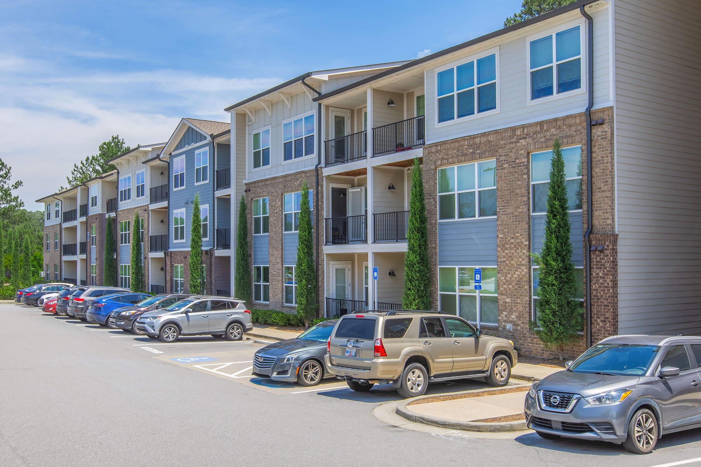A row of modern apartment buildings featuring various shades of exterior siding, with multiple vehicles parked in front. The scene is set under a clear blue sky with green trees in the background, showcasing a well-maintained residential area.