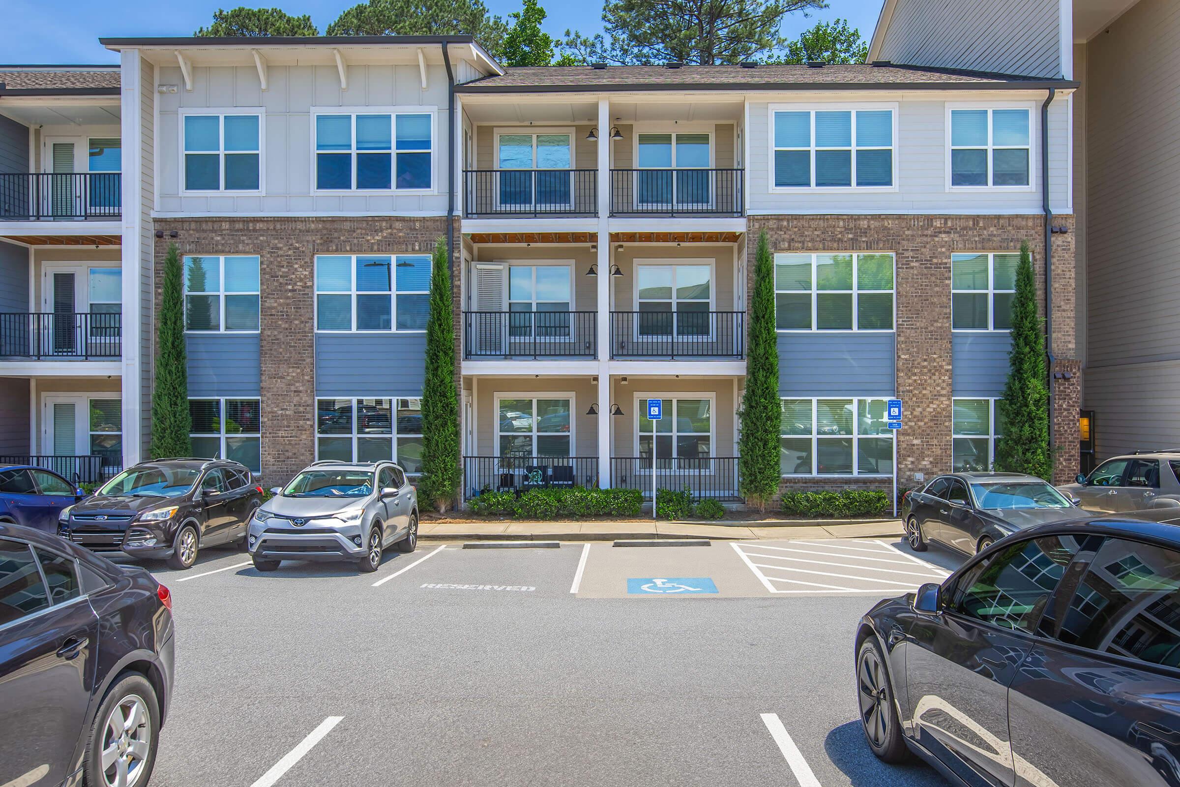 A multi-story residential building with several balconies, featuring large windows and a mix of brick and siding exterior. The scene includes several parked cars in the foreground, with designated parking spaces visible. Lush greenery is noted alongside the building, enhancing the welcoming atmosphere.