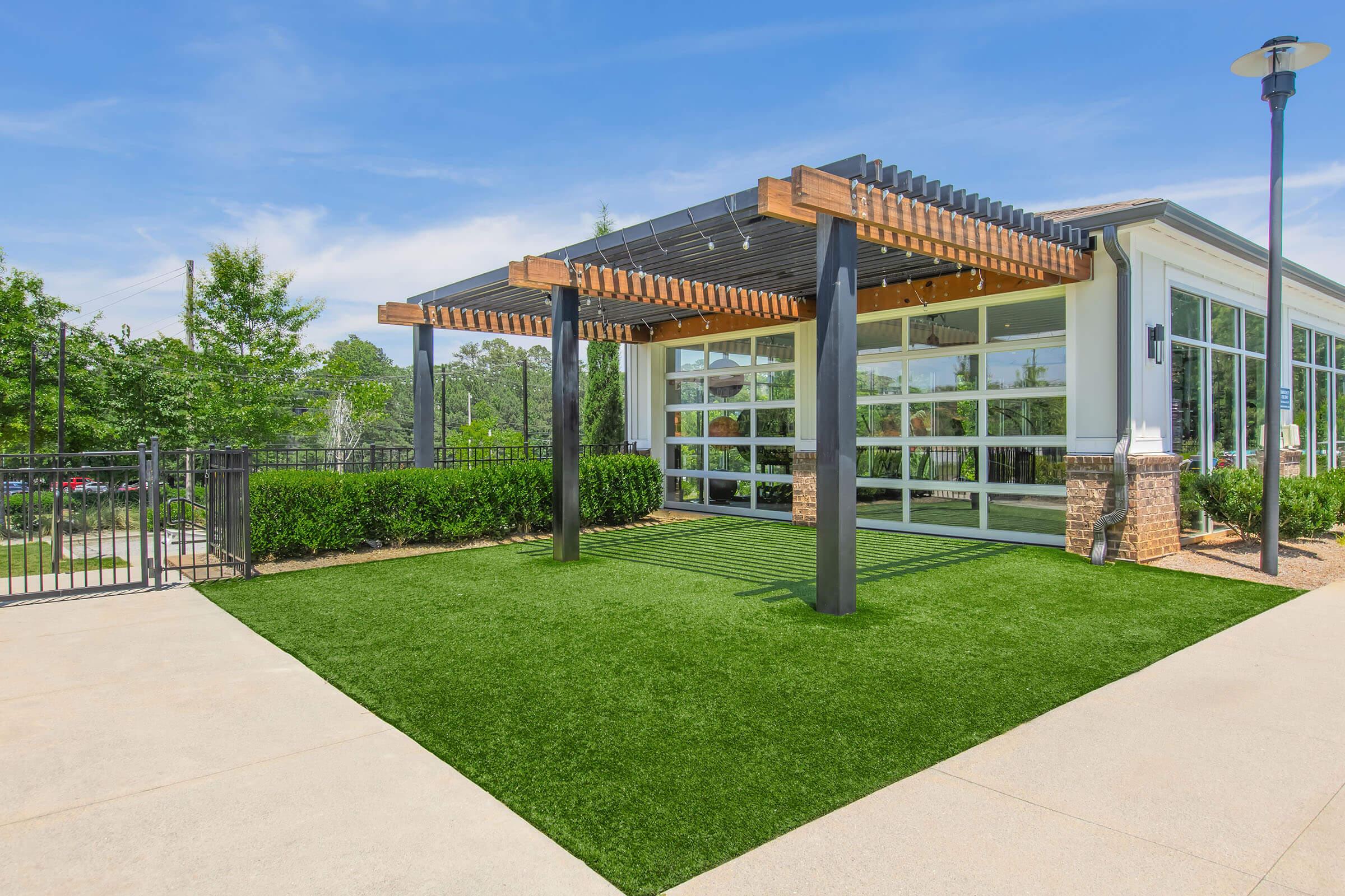 A modern outdoor space featuring a green lawn, a covered pergola, and large glass windows of a building. Lush trees and shrubs surround the area, creating a vibrant and inviting environment under a clear blue sky. The design combines natural elements with contemporary architecture.