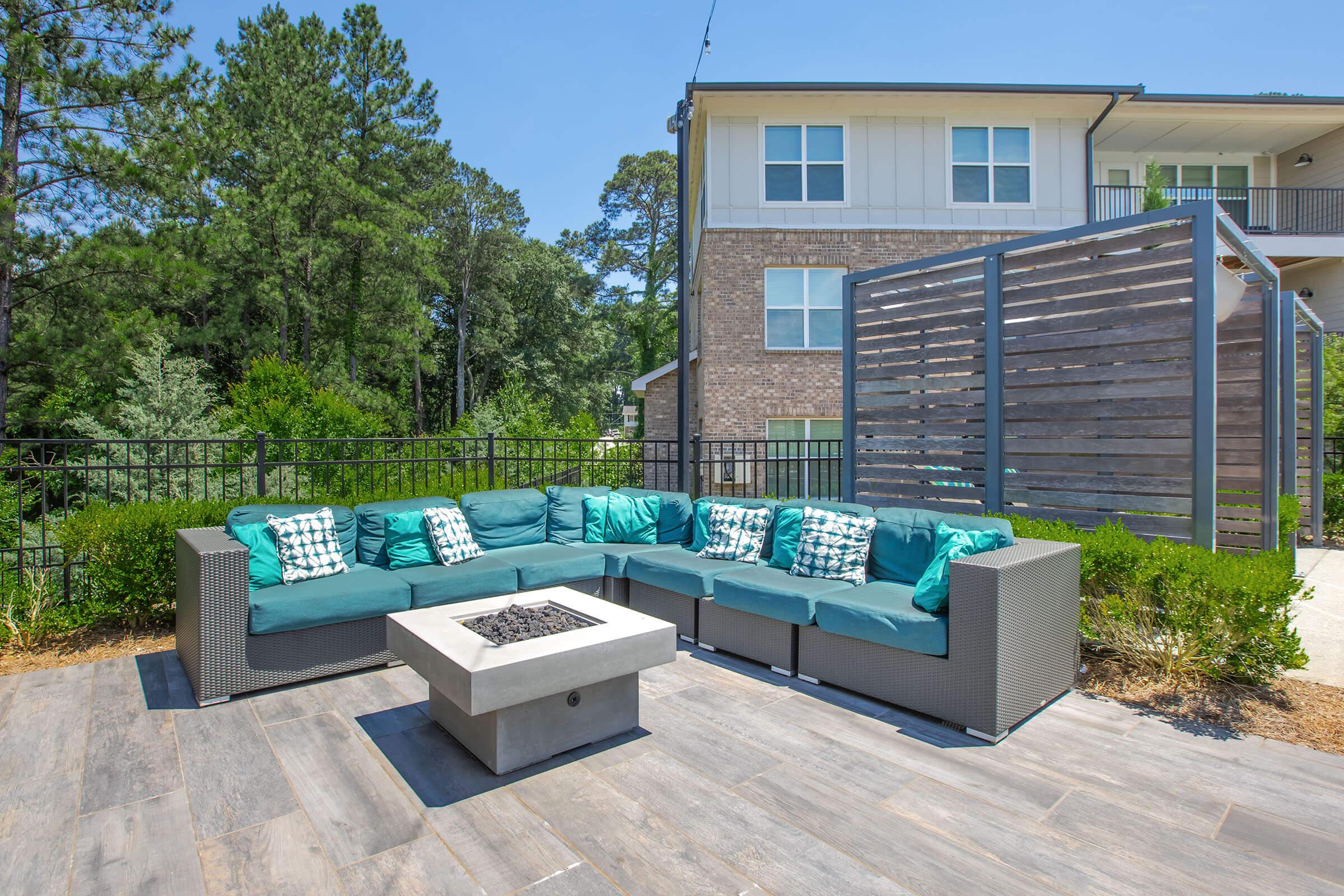 Outdoor seating area featuring a spacious sectional sofa with turquoise cushions and decorative pillows. In the center, there is a modern fire pit surrounded by a stone patio. The background includes trees and a two-story building with a balcony. The setting is bright and inviting, perfect for gatherings.