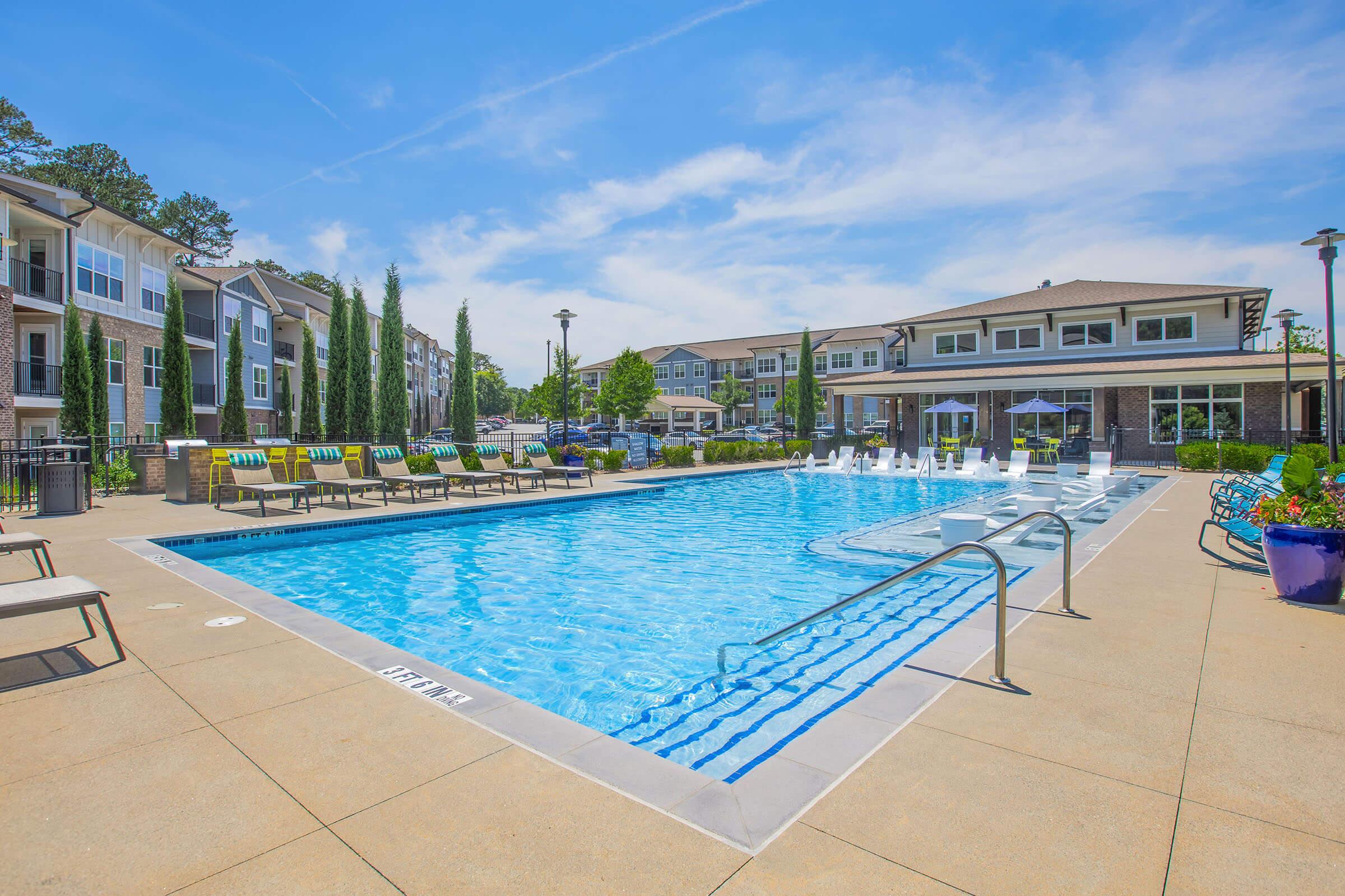A clear blue swimming pool surrounded by lounge chairs and umbrellas. In the background, there are modern apartment buildings with greenery and decorative plants, under a bright sky with scattered clouds. The scene is inviting and serene, ideal for relaxation and leisure.