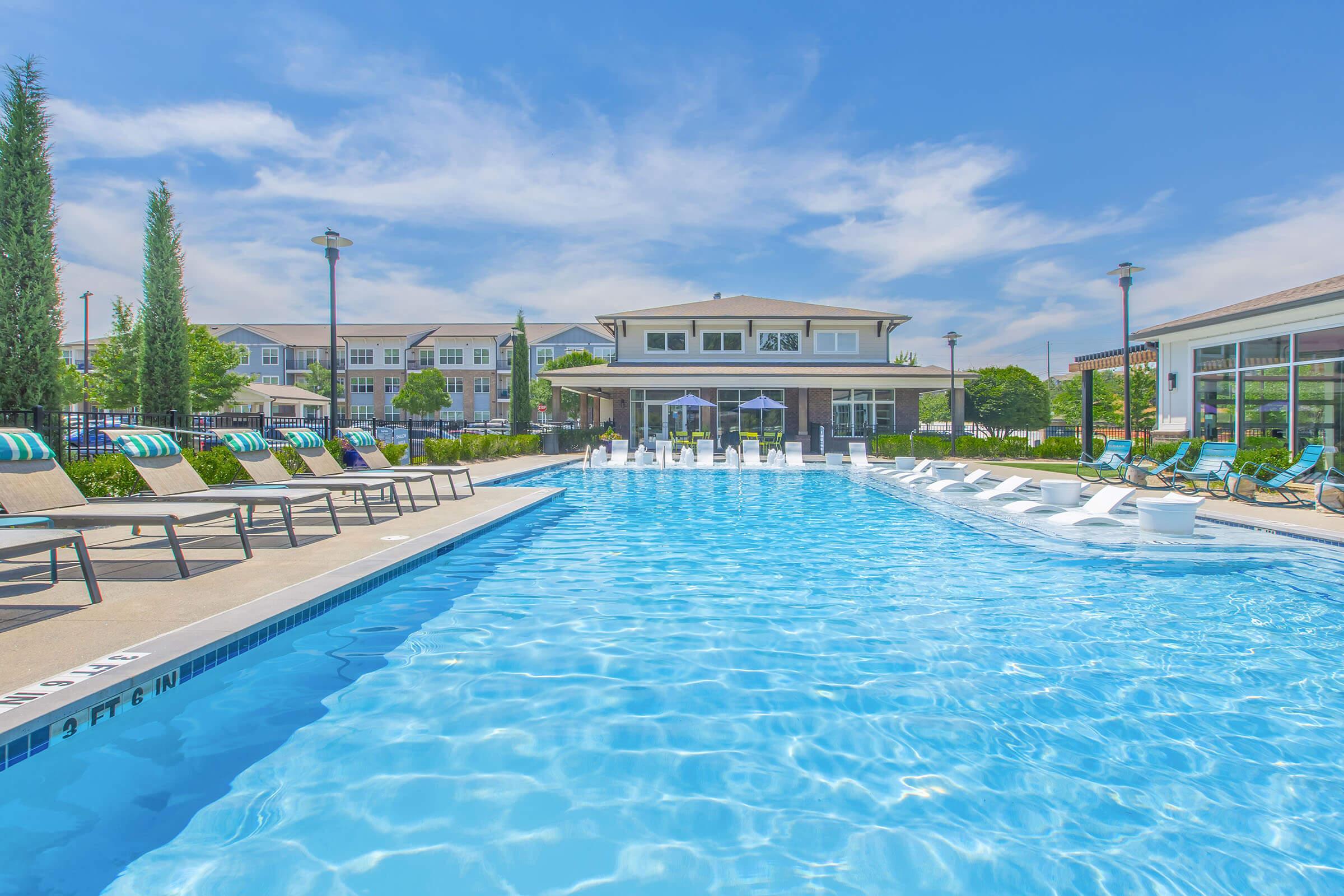 A bright blue swimming pool surrounded by lounge chairs and tall trees, with a two-story building in the background. Clear skies and fluffy clouds are visible overhead, creating a relaxing outdoor atmosphere.