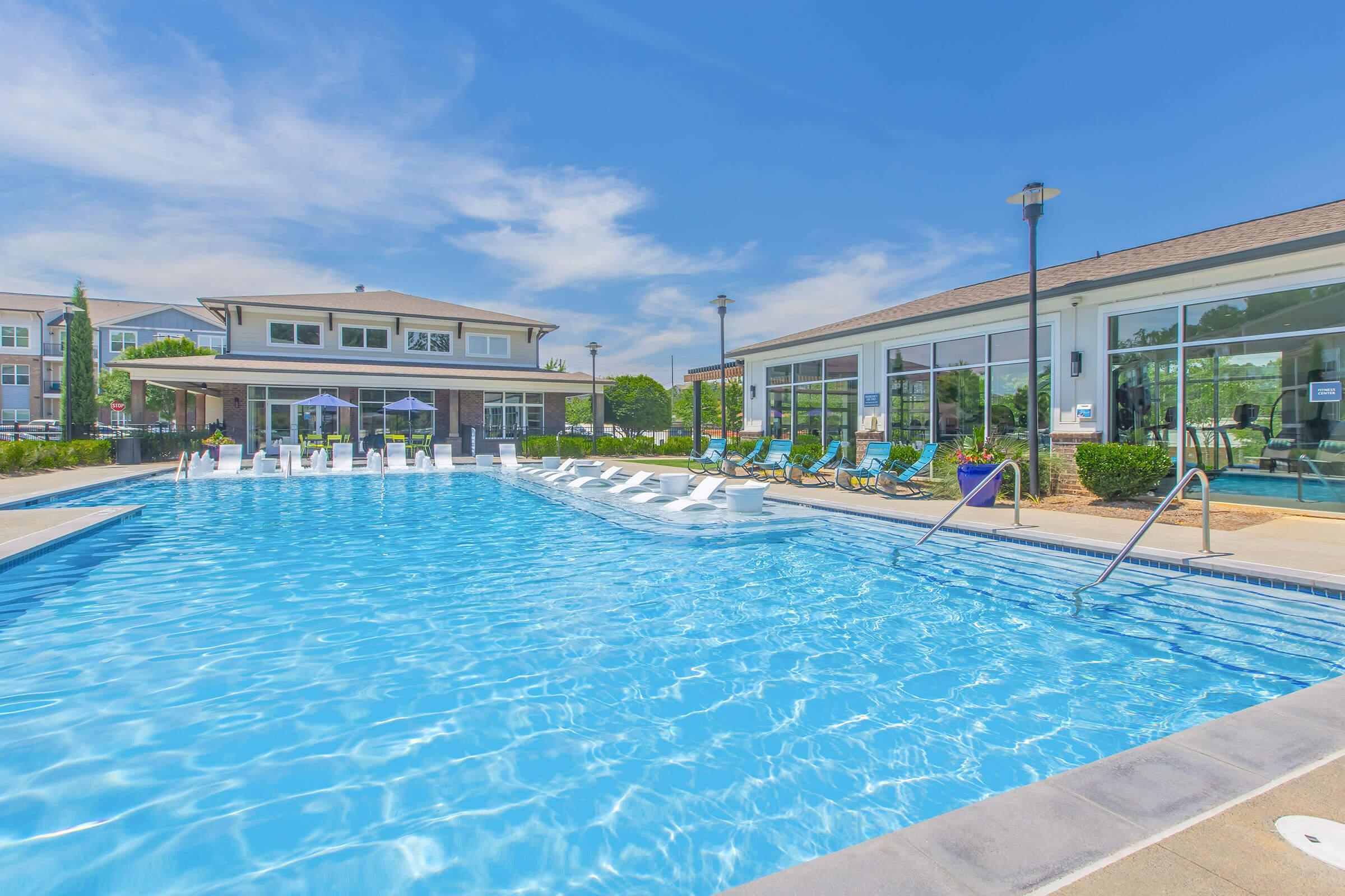A bright blue swimming pool surrounded by lounge chairs, with a modern building in the background. The sky is clear and sunny, creating a vibrant atmosphere. The pool area includes umbrellas and greenery, offering a relaxing setting for leisure and recreation.