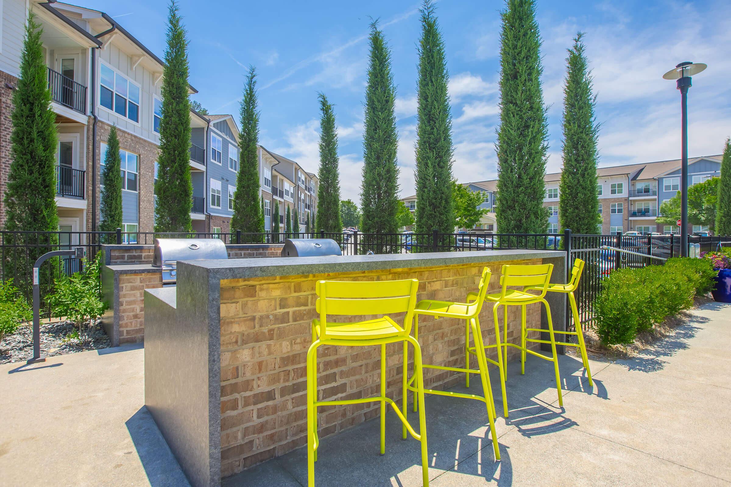 Outdoor barbecue area with a stone grill station and bright yellow bar stools. Lush, tall cypress trees line the space, creating a serene atmosphere. In the background, modern apartment buildings are visible under a clear blue sky. The scene conveys a relaxed, inviting outdoor living environment.